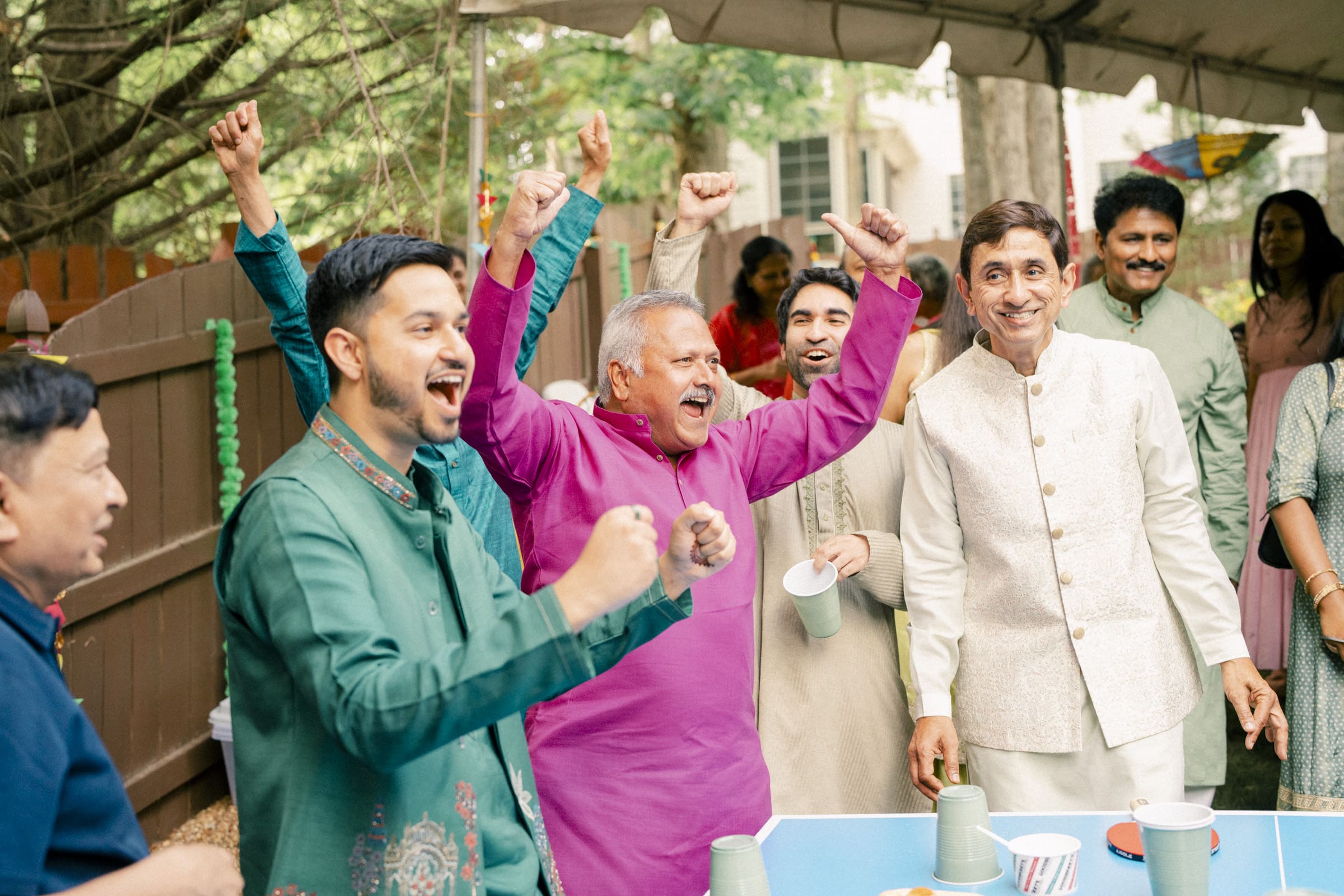 A group of men in traditional South Asian attire cheer and smile excitedly around a table outdoors, appearing to celebrate or play a game at a festive gathering.
