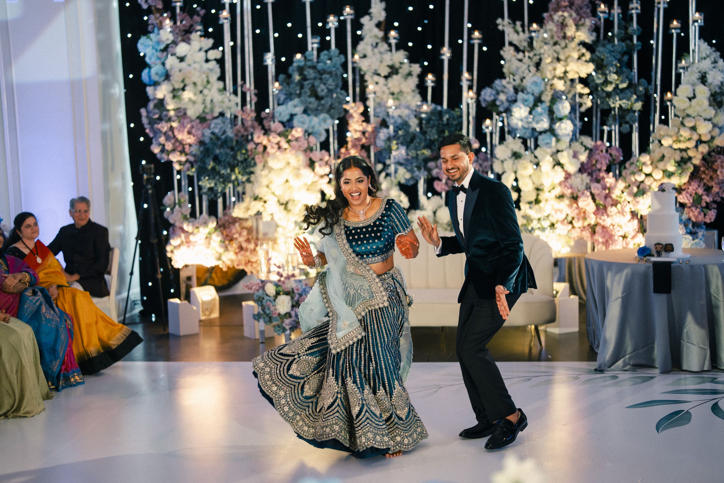 A smiling couple in formal attire dance together on an elegant stage adorned with towering white floral arrangements at Donovan Manor.
