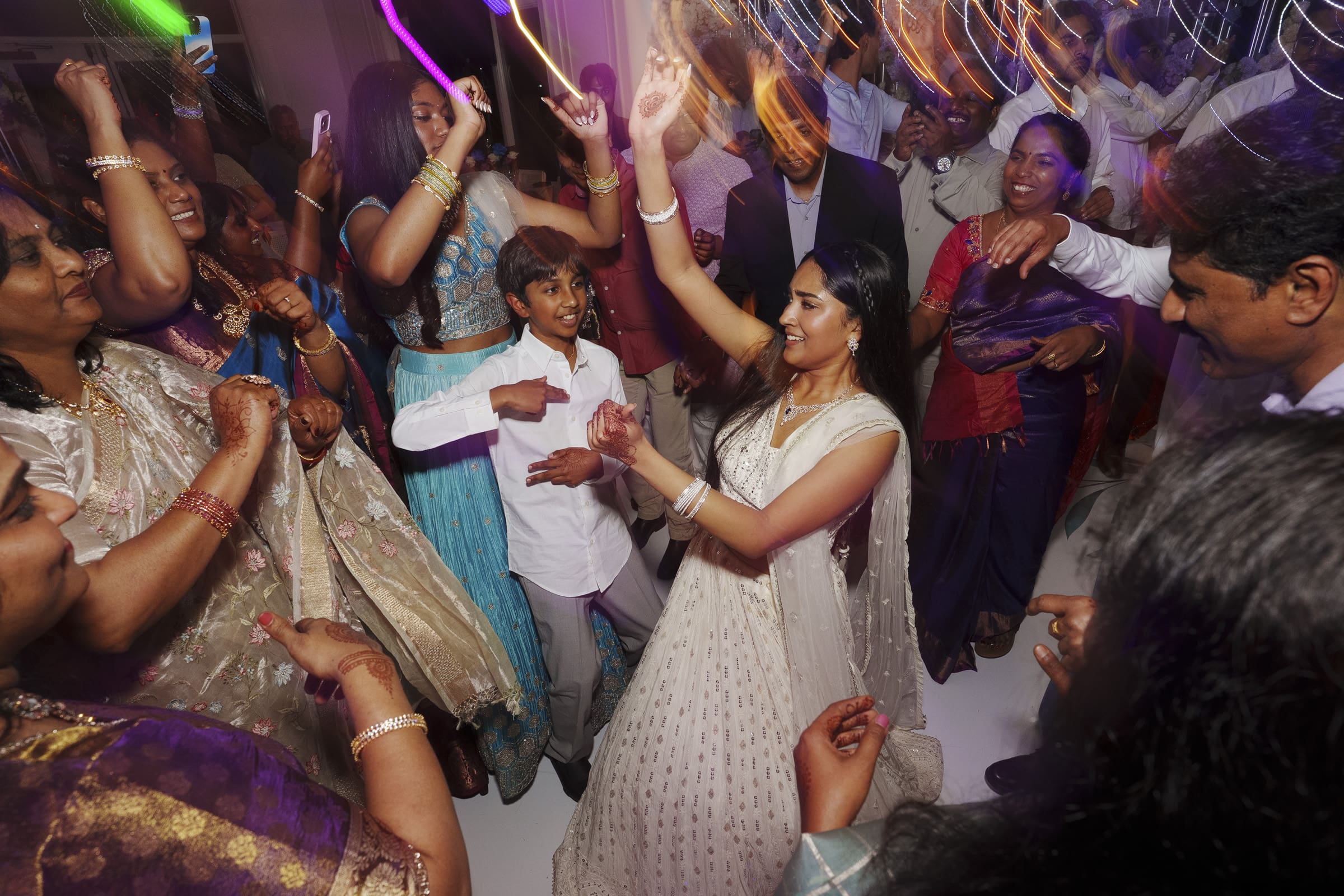 A crowd of wedding guests in vibrant traditional Indian clothing dance together with raised hands on a packed dance floor.