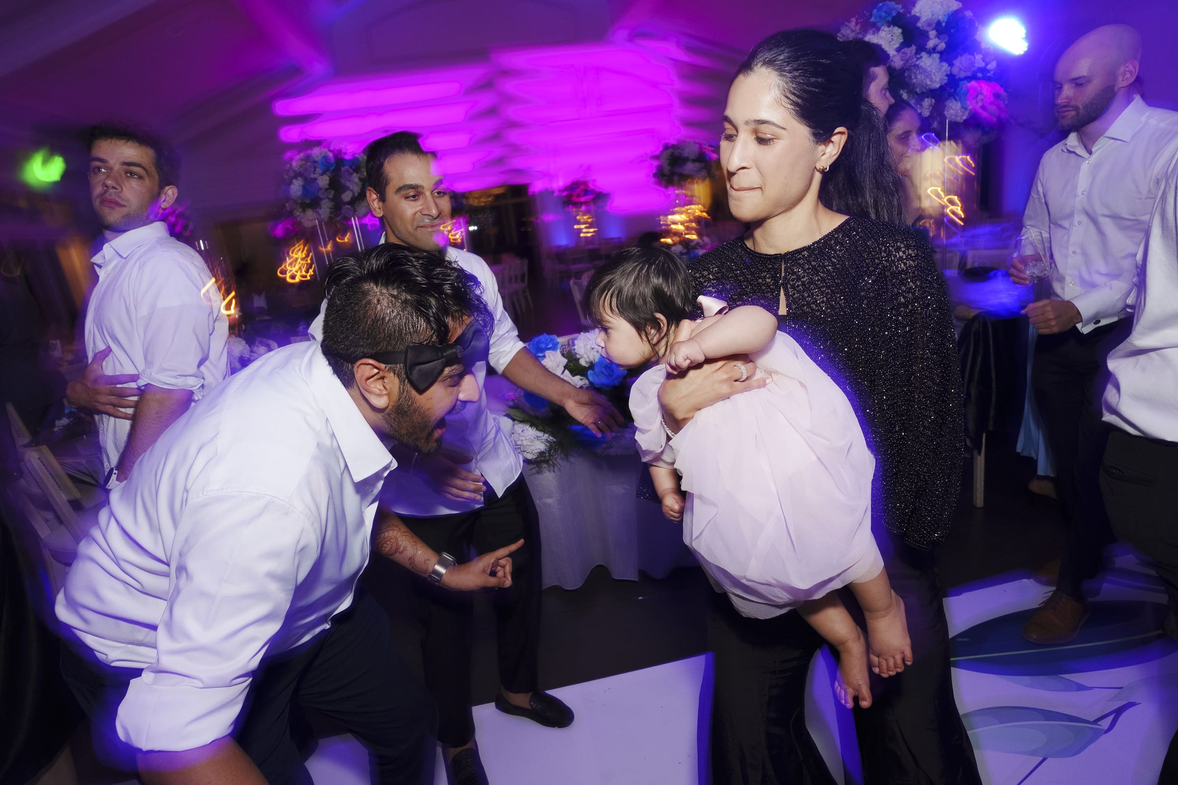 A guest playfully interacts with a baby on a vibrant dance floor surrounded by family under purple and blue reception party lighting.