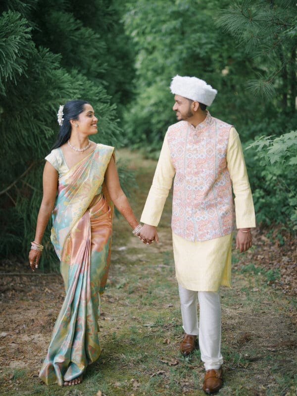 A couple in traditional Indian attire walks hand-in-hand along a wooded path. The woman wears a colorful saree; the man wears a pastel kurta with a patterned vest and white cap. They smile warmly at each other, surrounded by greenery.