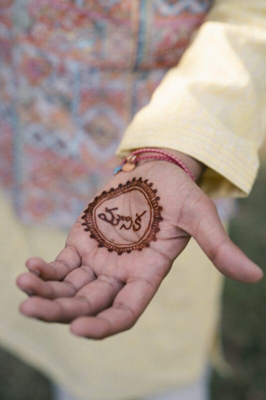 A close-up of an outstretched hand with henna art and Telugu script on the palm. The person is wearing a yellow, patterned traditional outfit and a red thread bracelet. The background is blurred greenery.