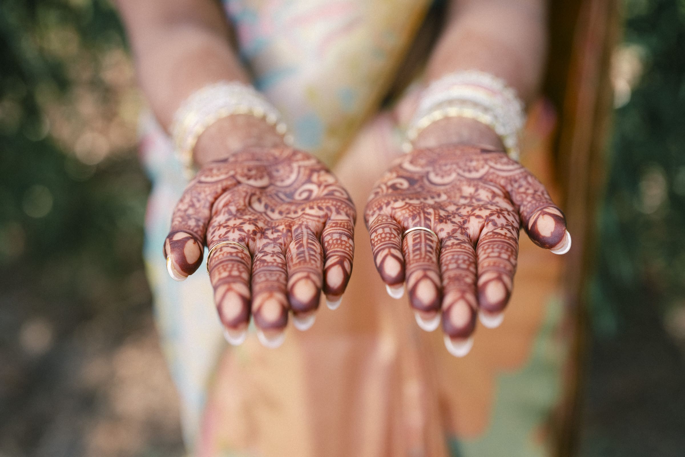 A person wearing intricate bangles holds out their hands, showing detailed henna designs covering their palms and fingers, with a colorful, blurred background.