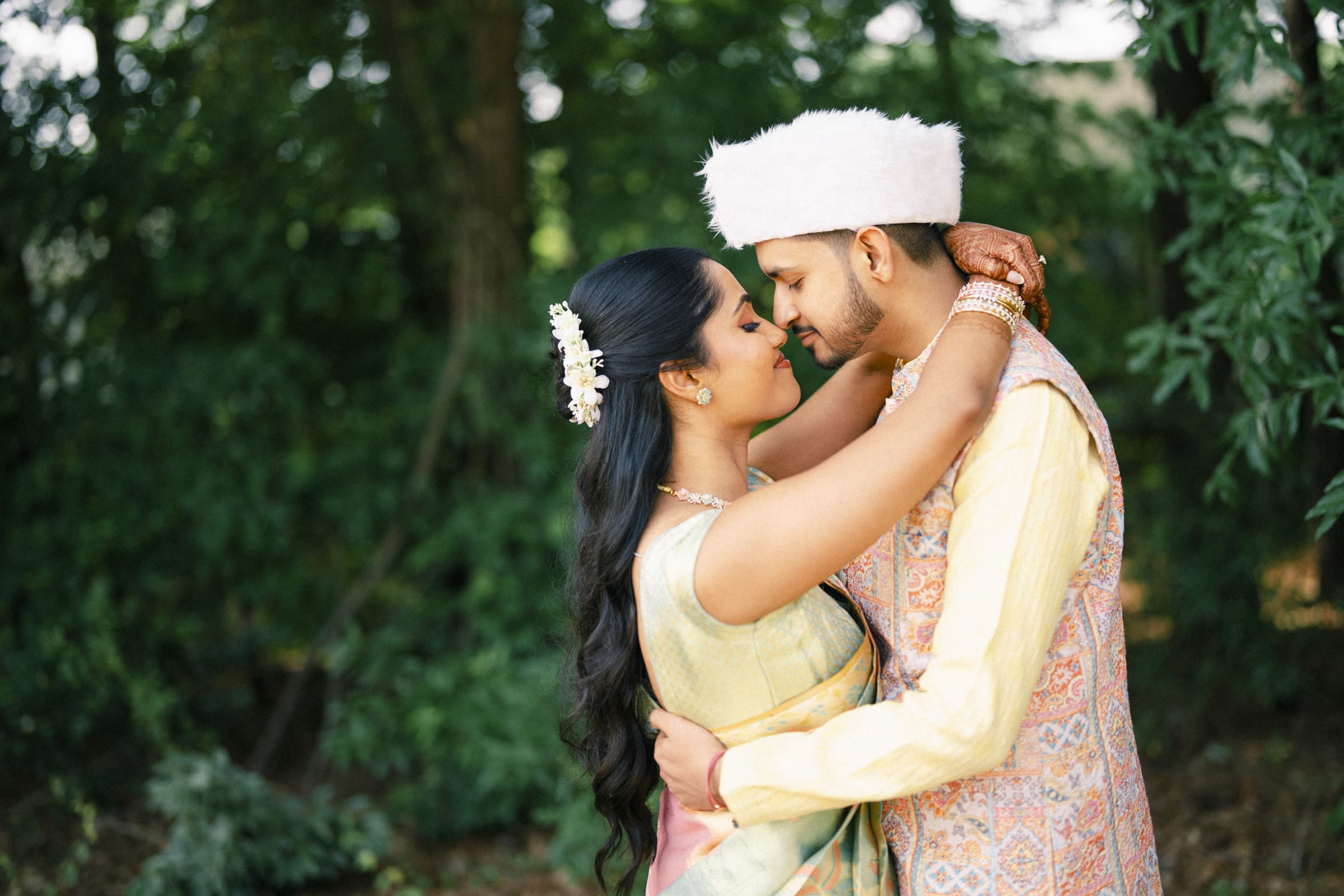 A couple dressed in traditional South Asian wedding attire lovingly embrace outdoors, surrounded by greenery. The woman has long dark hair adorned with white flowers, and the man wears a white fur hat and embroidered vest.