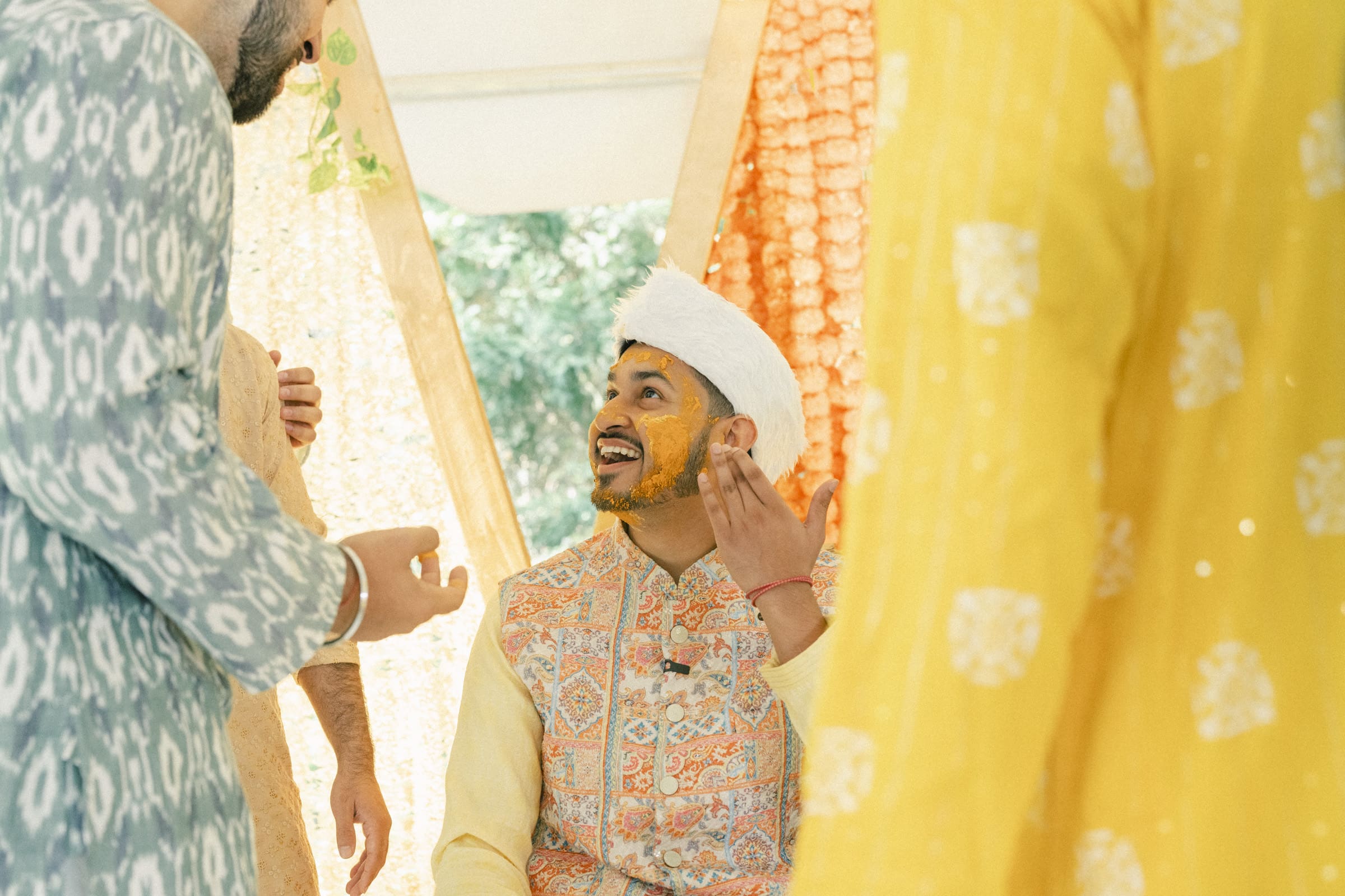 A man dressed in traditional attire with turmeric on his face smiles as he participates in a pre-wedding ceremony, surrounded by others in colorful clothing under festive decorations.