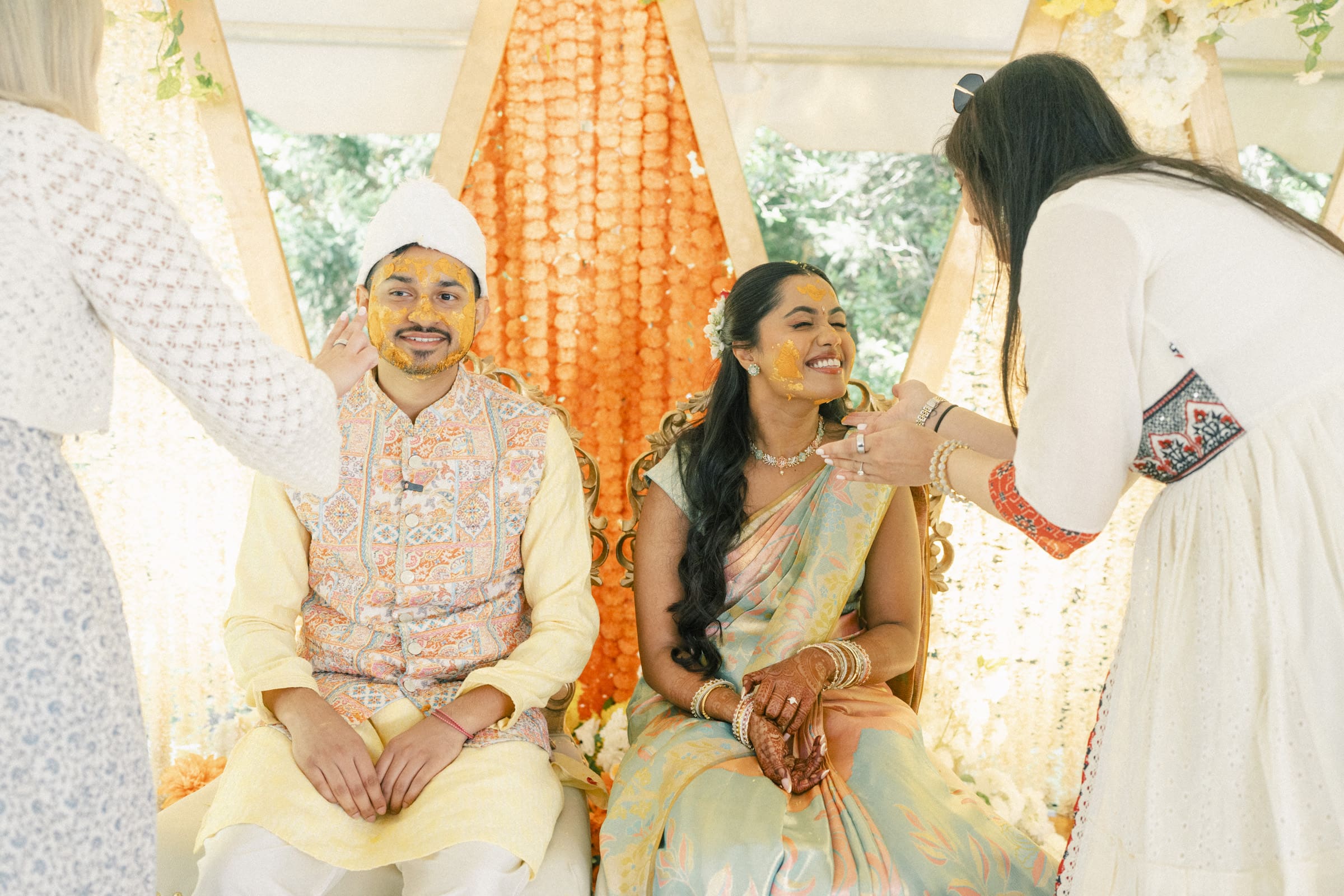 A couple in traditional attire sit side by side smiling while two women apply turmeric paste to their faces during a pre-wedding ceremony. Orange and white floral decorations hang behind them.