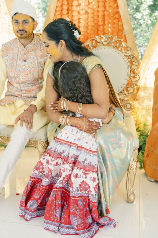 A woman in a pastel traditional outfit sits on an ornate chair, hugging a young girl in a colorful dress. Both have turmeric on their faces, suggesting a pre-wedding ceremony. A man in traditional attire sits beside them.