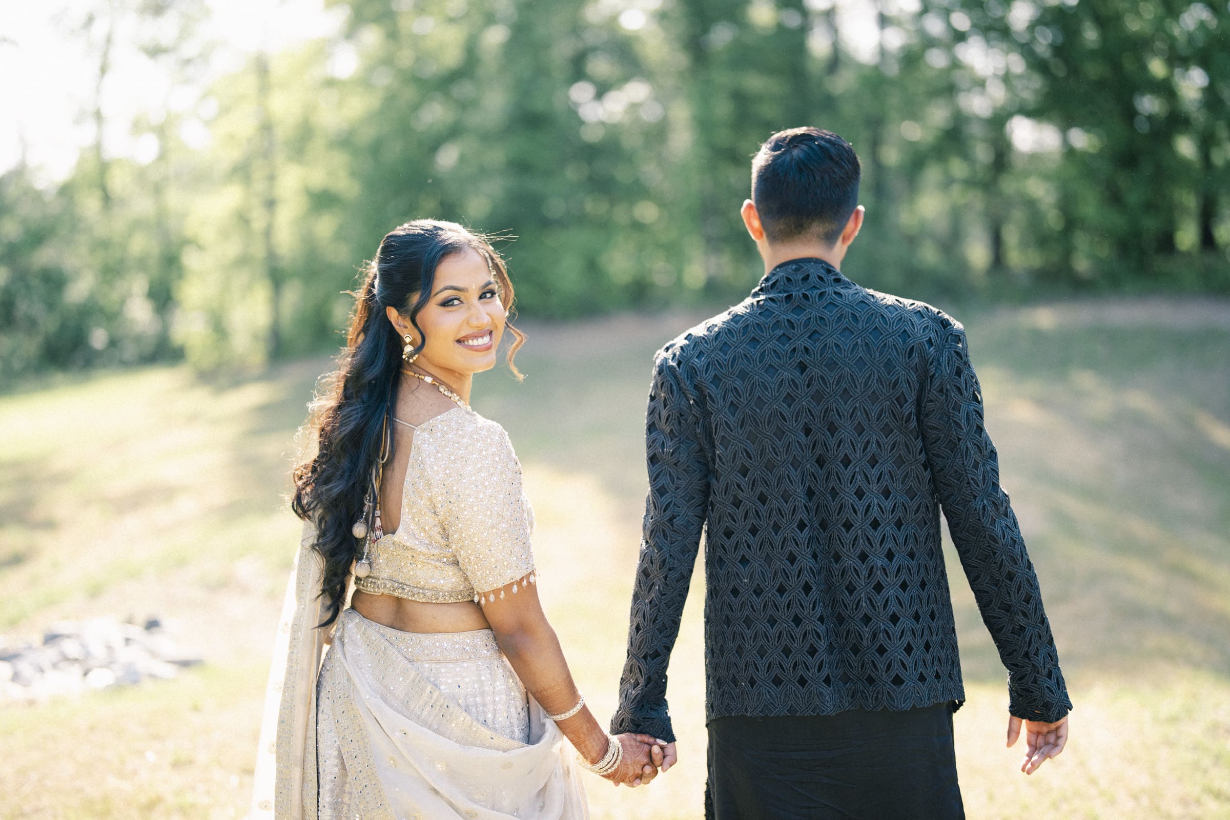 A smiling woman in traditional South Asian attire holds hands with a man in a black patterned outfit as they walk outdoors on a sunny day, surrounded by greenery at a Raleigh Indian wedding by Joe Payne Photography..