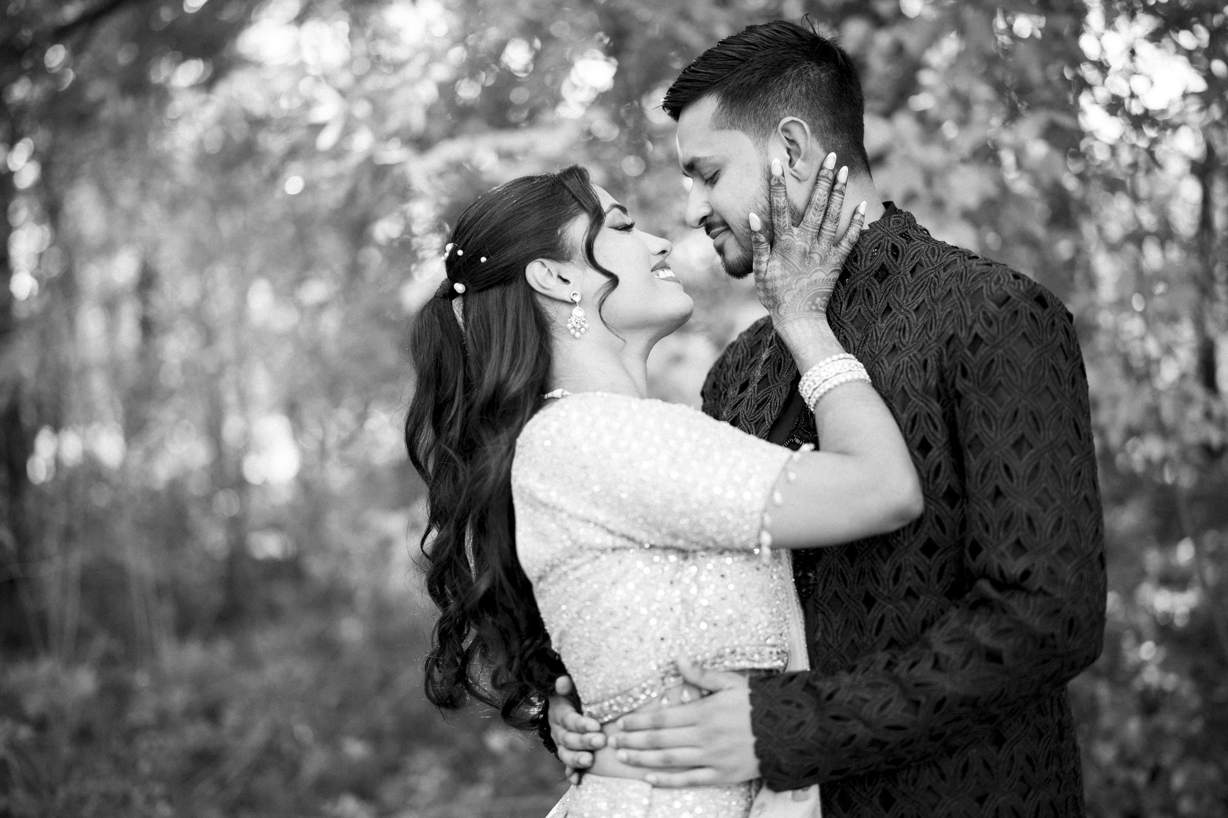 A couple in traditional attire embrace and smile lovingly at each other in an outdoor setting, surrounded by blurred trees and foliage. The woman gently holds the mans face while he smiles at her at a Raleigh Indian wedding by Joe Payne Photography..
