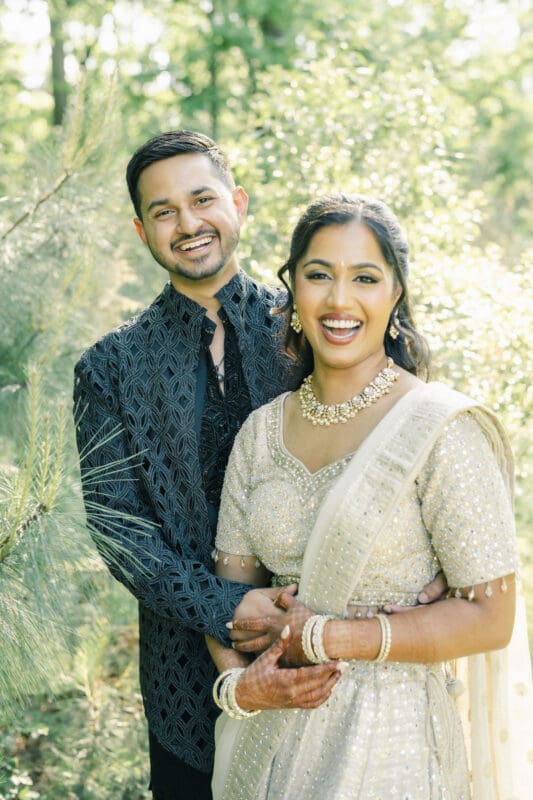 A smiling couple in traditional South Asian attire stands outdoors among greenery. The woman wears a cream and gold sari with jewelry and henna on her hands; the man stands behind her in a dark embroidered kurta at a Raleigh Indian wedding by Joe Payne Photography..