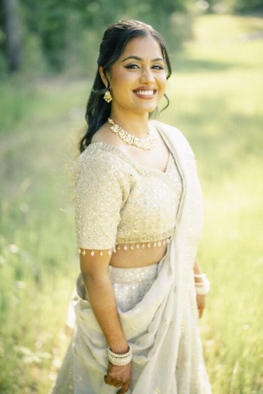 A woman in a light-colored, embellished saree stands outdoors, smiling warmly. She wears jewelry, including a necklace and bangles, with her hair styled in soft waves. Sunlight brightens the natural, green background at a Raleigh Indian wedding by Joe Payne Photography..
