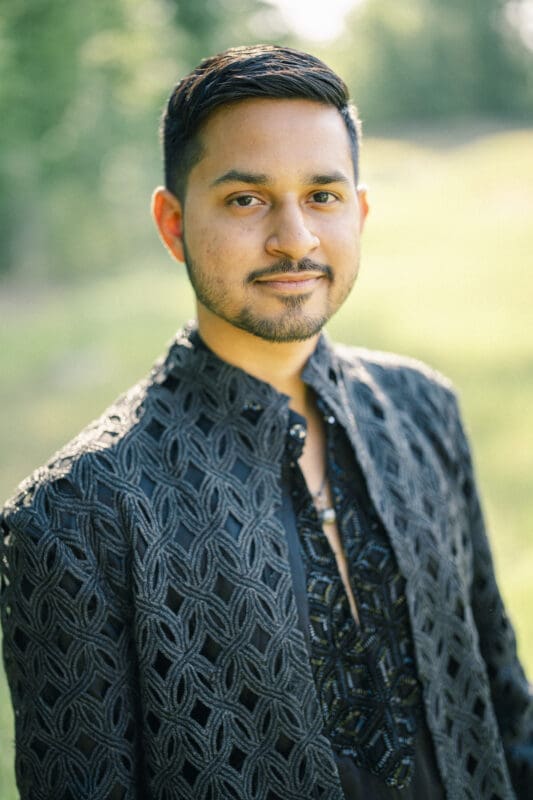 A man with short dark hair and a trimmed beard stands outdoors, wearing an ornate black embroidered jacket. The background is blurred greenery, and sunlight gently illuminates his face at a Raleigh Indian wedding by Joe Payne Photography..