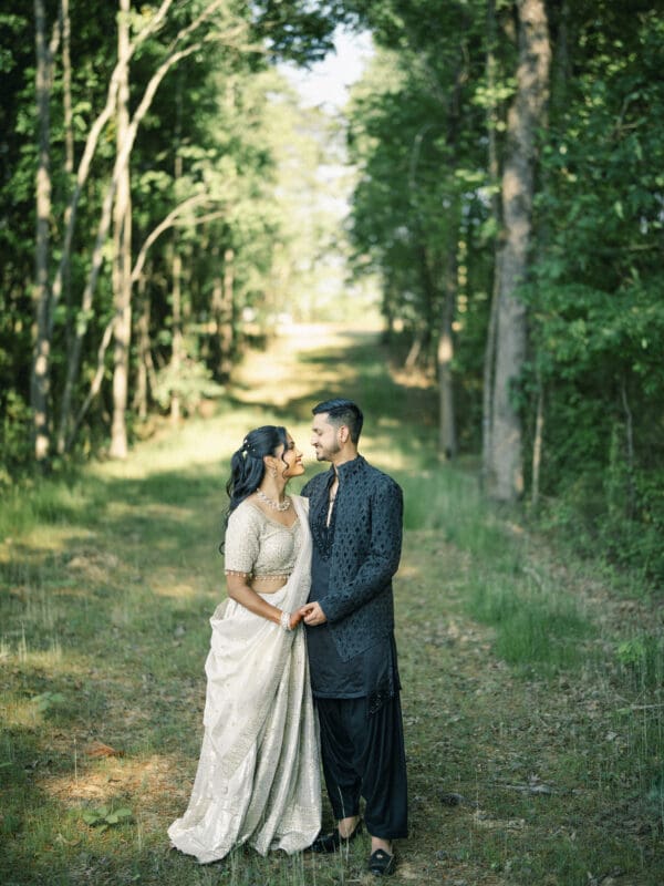 A couple stands closely together on a sunlit forest path, smiling at each other. The woman wears a cream and gold traditional outfit, and the man wears a black kurta with matching pants. Green trees line both sides of the path at a Raleigh Indian wedding by Joe Payne Photography..