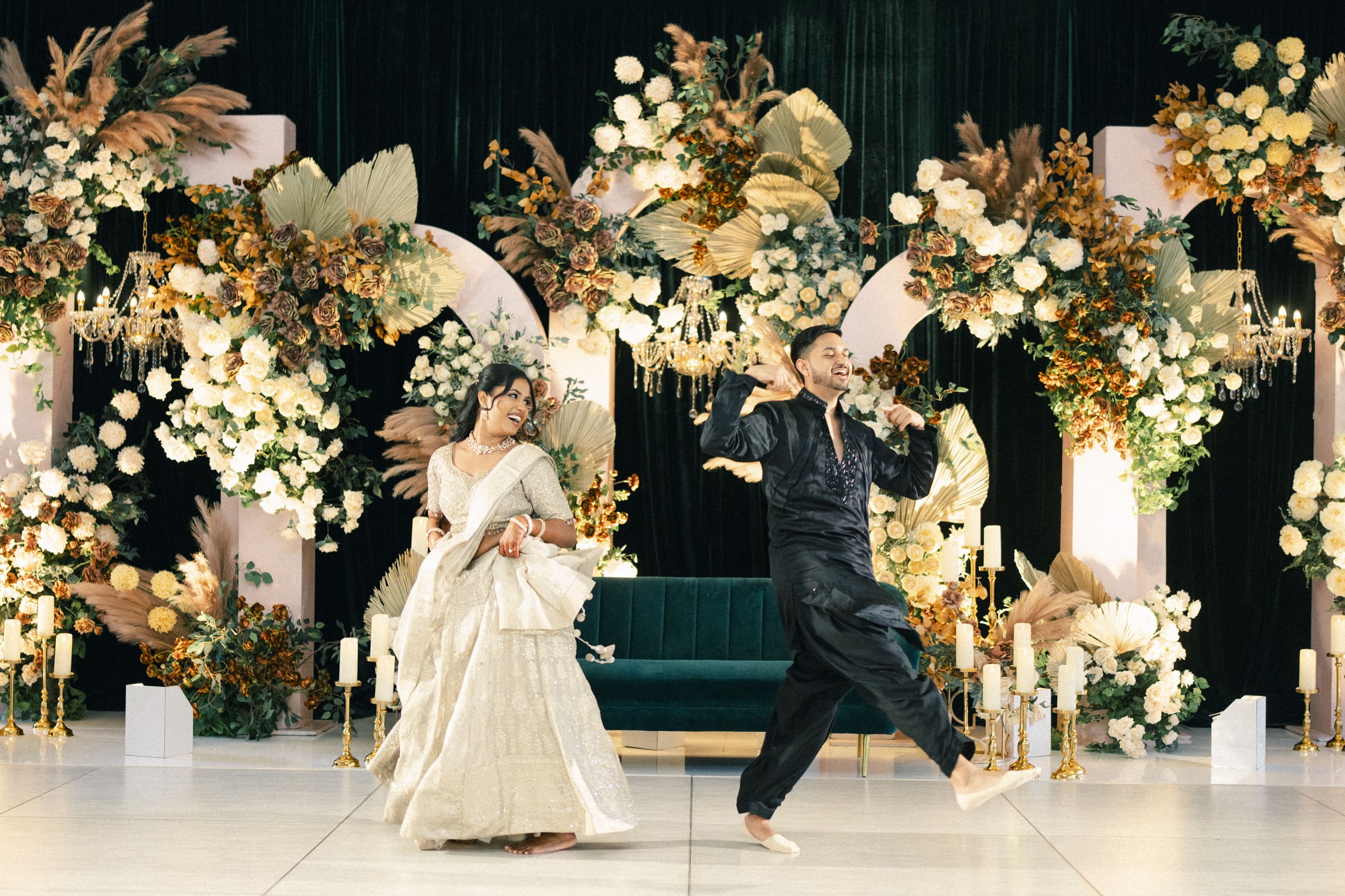 A joyful couple in traditional South Asian attire dances in front of a lush floral backdrop with gold accents, chandeliers, and a green velvet sofa at at a Raleigh Indian wedding by Joe Payne Photography.