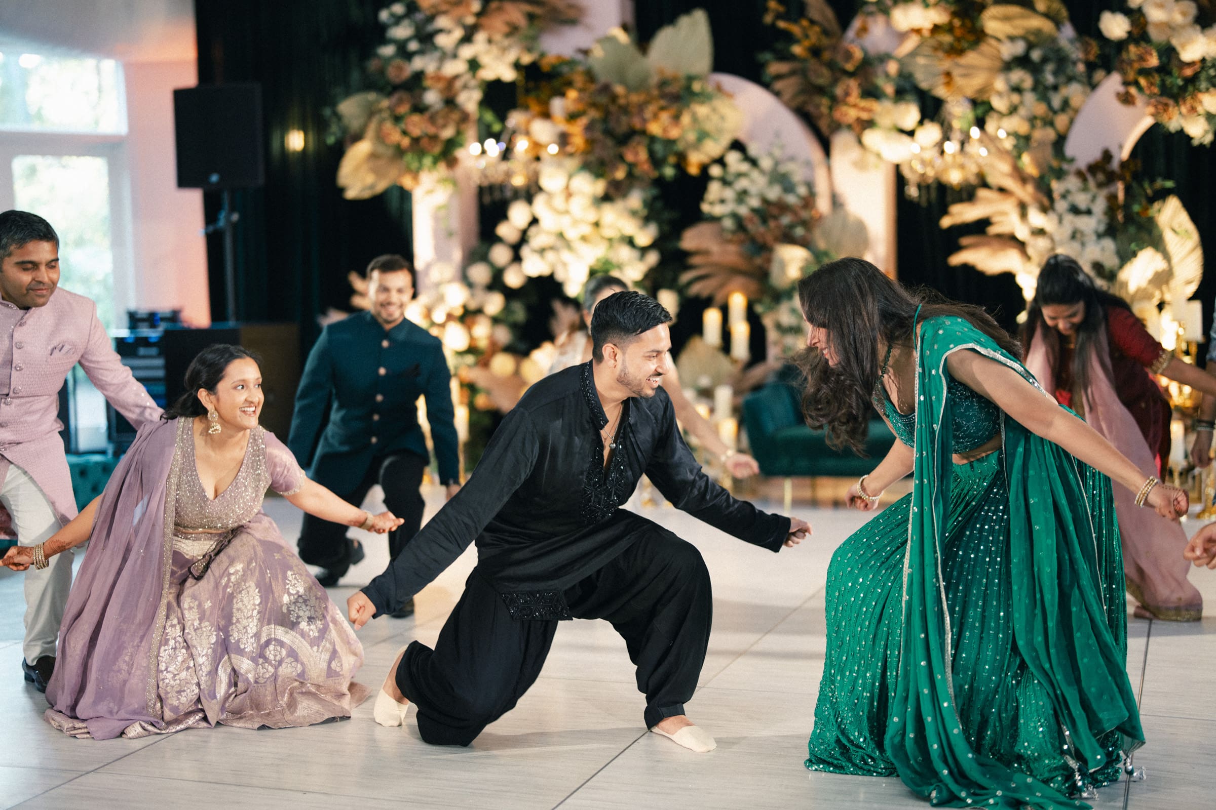 A group of people in colorful traditional Indian attire energetically dance together on a decorated indoor floor, smiling and enjoying the festive celebration.