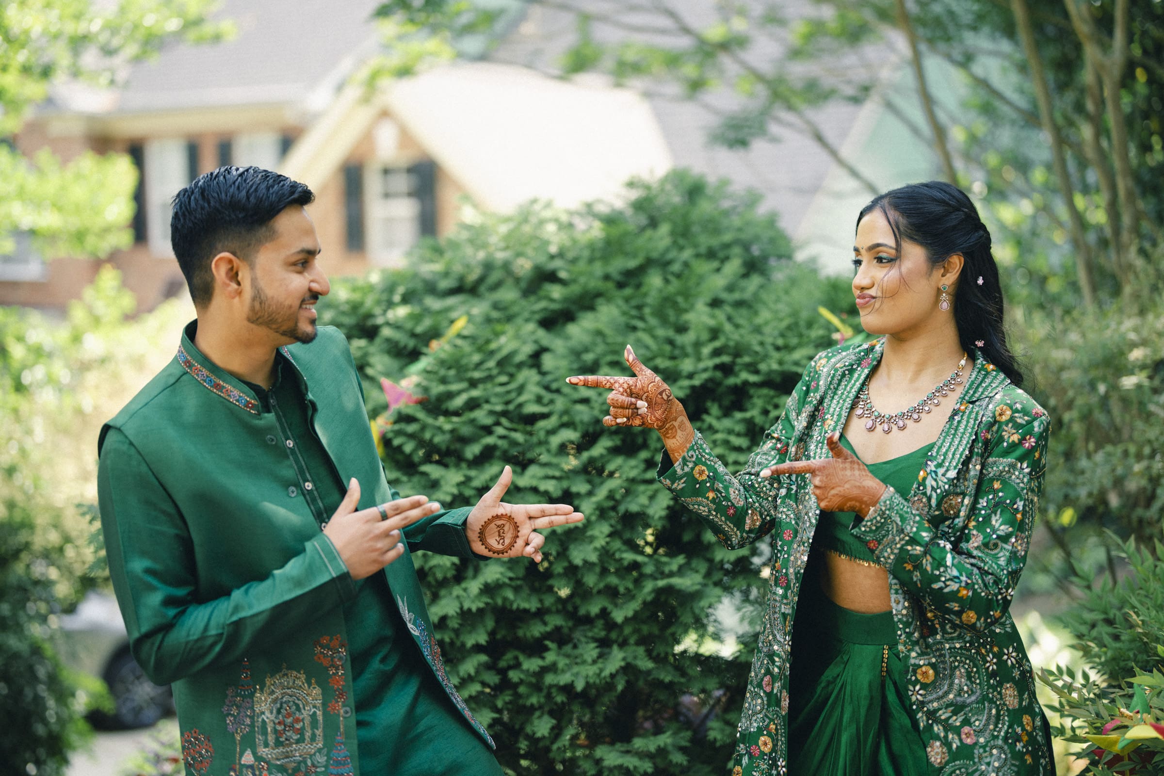 A man and woman in coordinated green traditional outfits smile and playfully point at each other outdoors, surrounded by lush greenery. The woman has henna designs on her hands and both look happy and engaged.