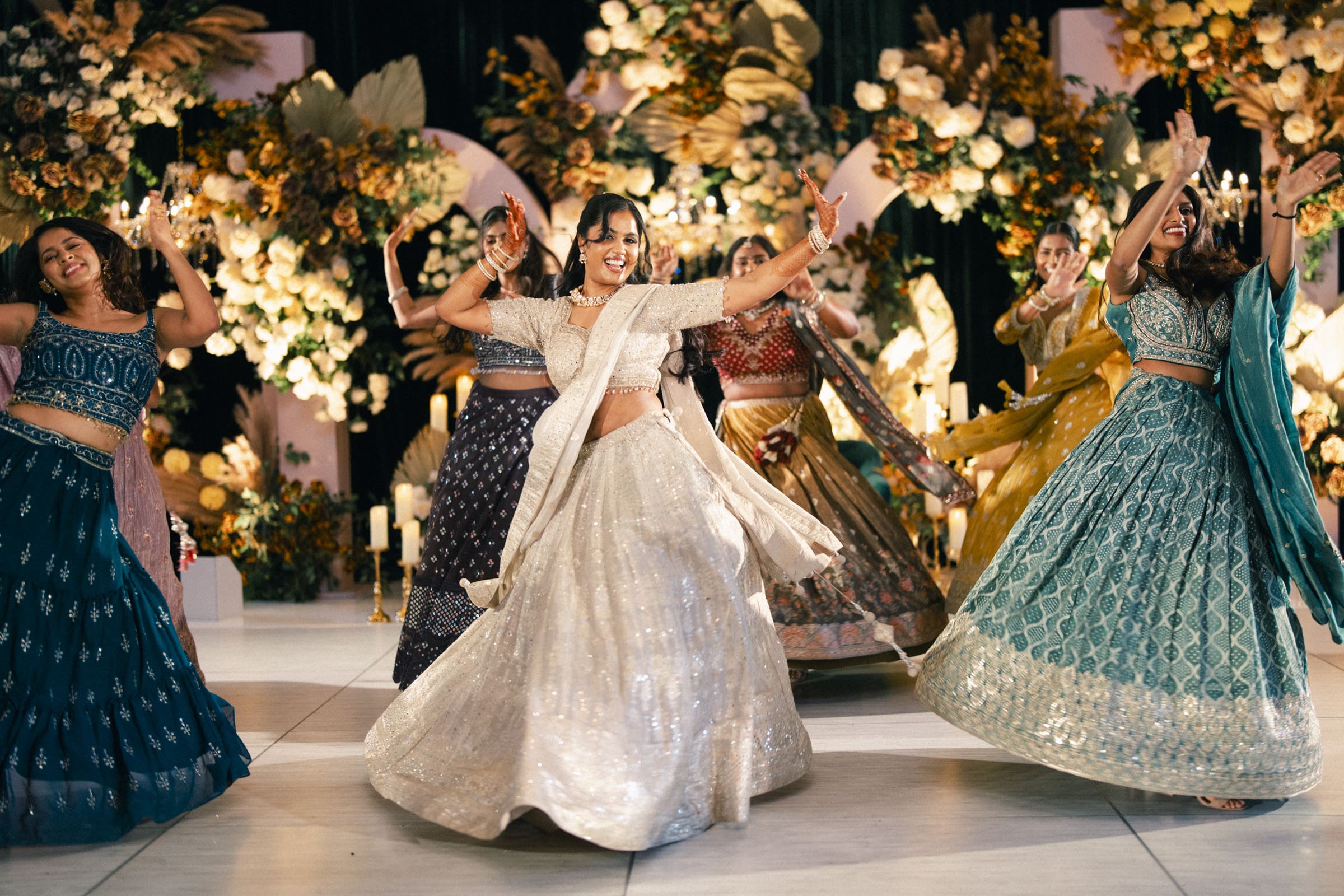 A lively group of wedding guests in colorful traditional Indian attire dance joyfully on an indoor stage, surrounded by elegant floral arrangements and dramatic stage lighting at a Raleigh Indian wedding by Joe Payne Photography.