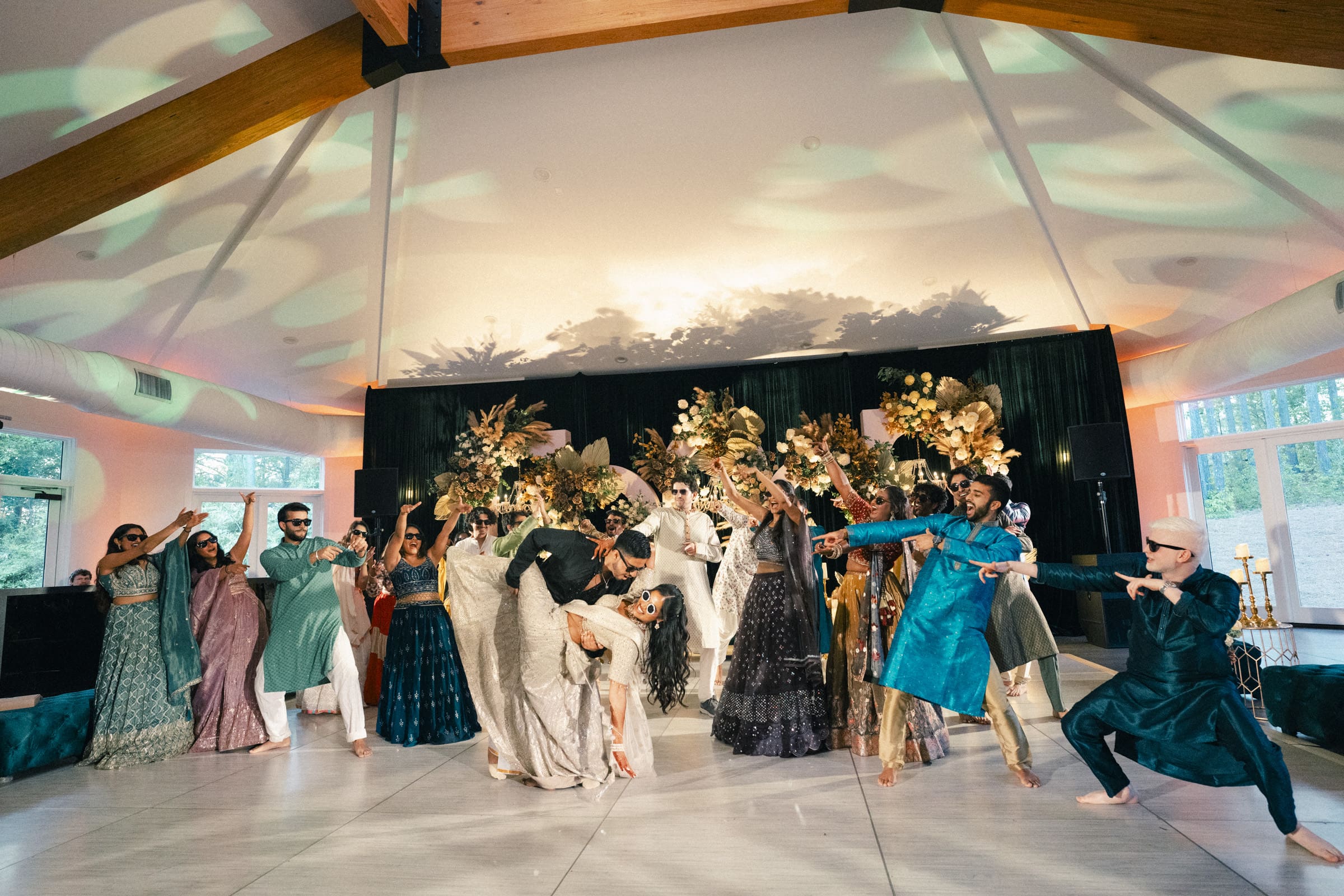 A group of people dressed in colorful traditional South Asian attire dance joyfully on a decorated indoor stage with floral arrangements and dramatic lighting.