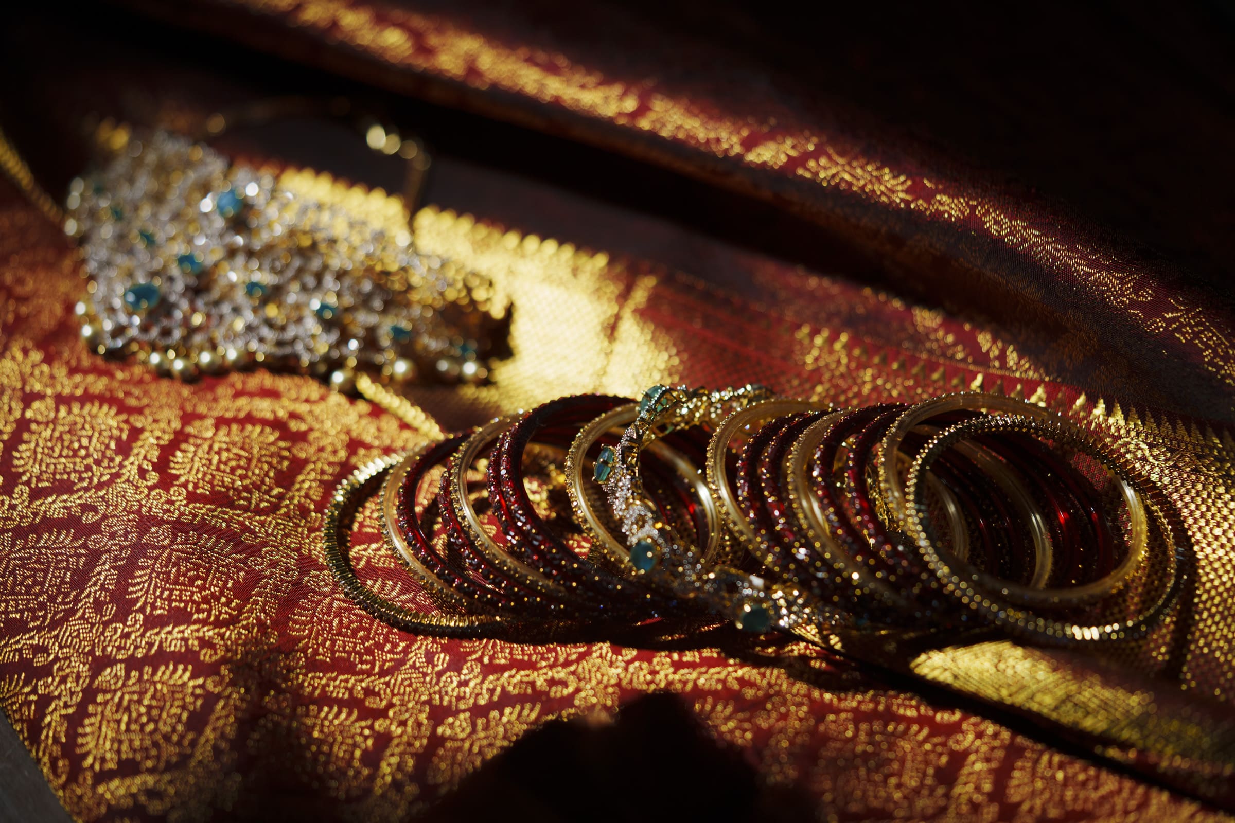 Gold and red bangles and a jeweled necklace rest on ornate red and gold fabric, illuminated by warm light.