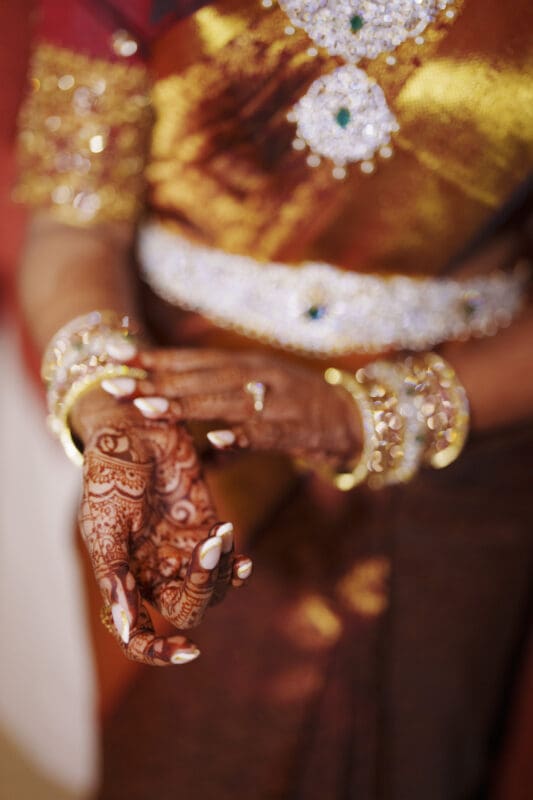 A woman wearing a gold and red saree with intricate jewelry and bangles displays her hands adorned with detailed henna designs and manicured nails.