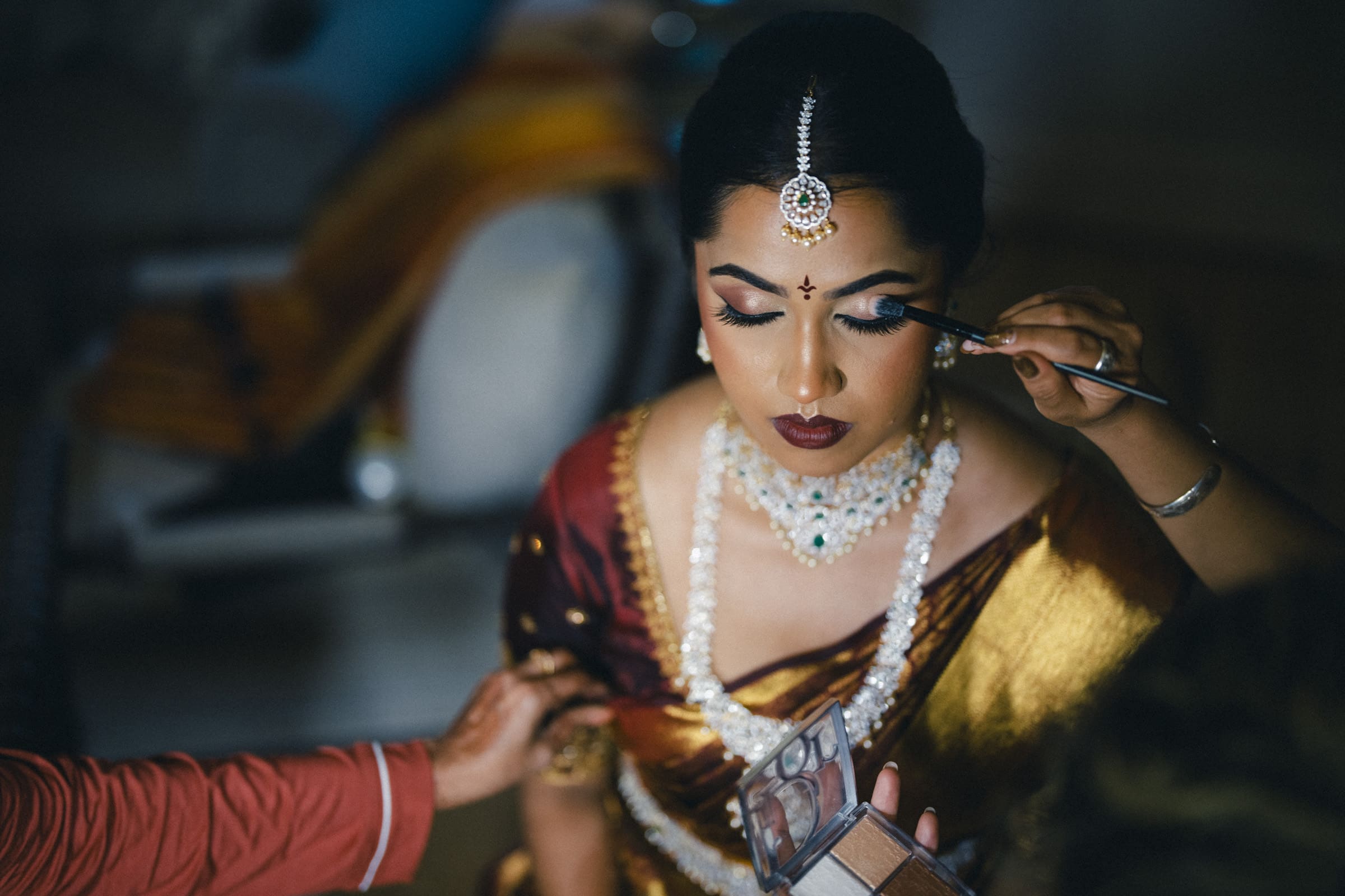 A woman in traditional Indian attire and jewelry sits with her eyes closed as a makeup artist applies eyeshadow. She wears a gold and maroon sari, elaborate necklace, earrings, and a bindi on her forehead.
