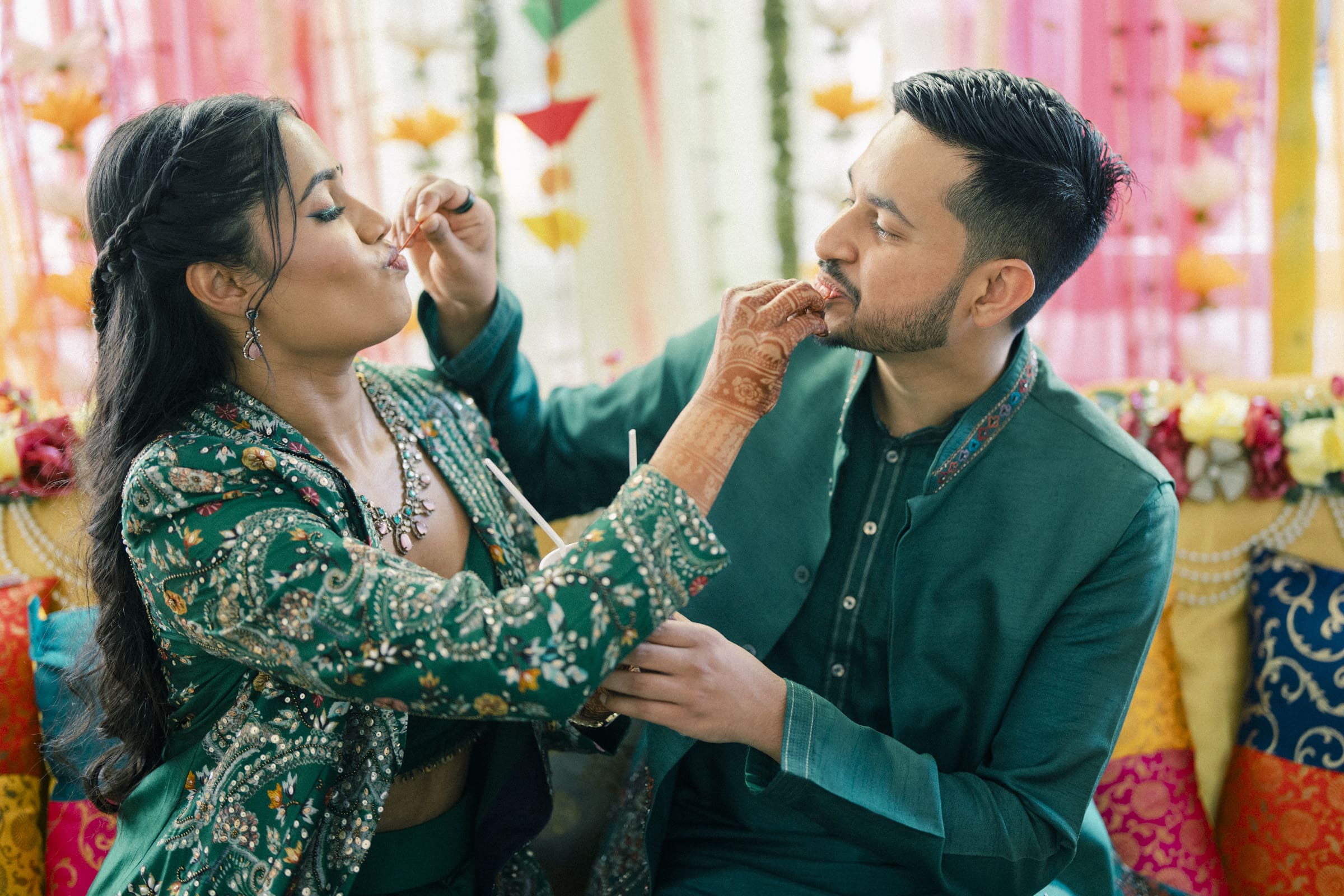 A couple dressed in traditional green outfits feed each other sweets, smiling and enjoying a festive celebration. Brightly colored decorations and pillows are in the background.