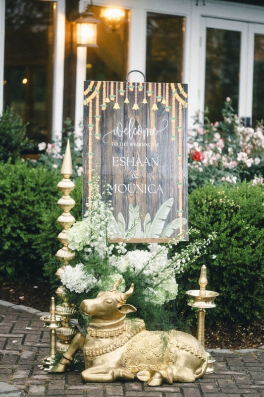 A welcome sign for a wedding, decorated with gold accents, white flowers, and a gold cow statue, stands in front of lush greenery and a building with white framed windows.