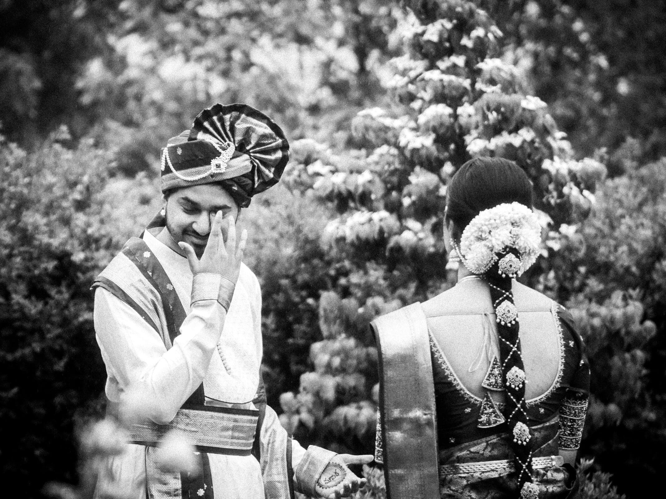 A groom in traditional Indian attire smiles and touches his face while standing beside a bride, who has her back to the camera and wears an ornate saree with flowers in her hair. Both are outdoors, surrounded by greenery.