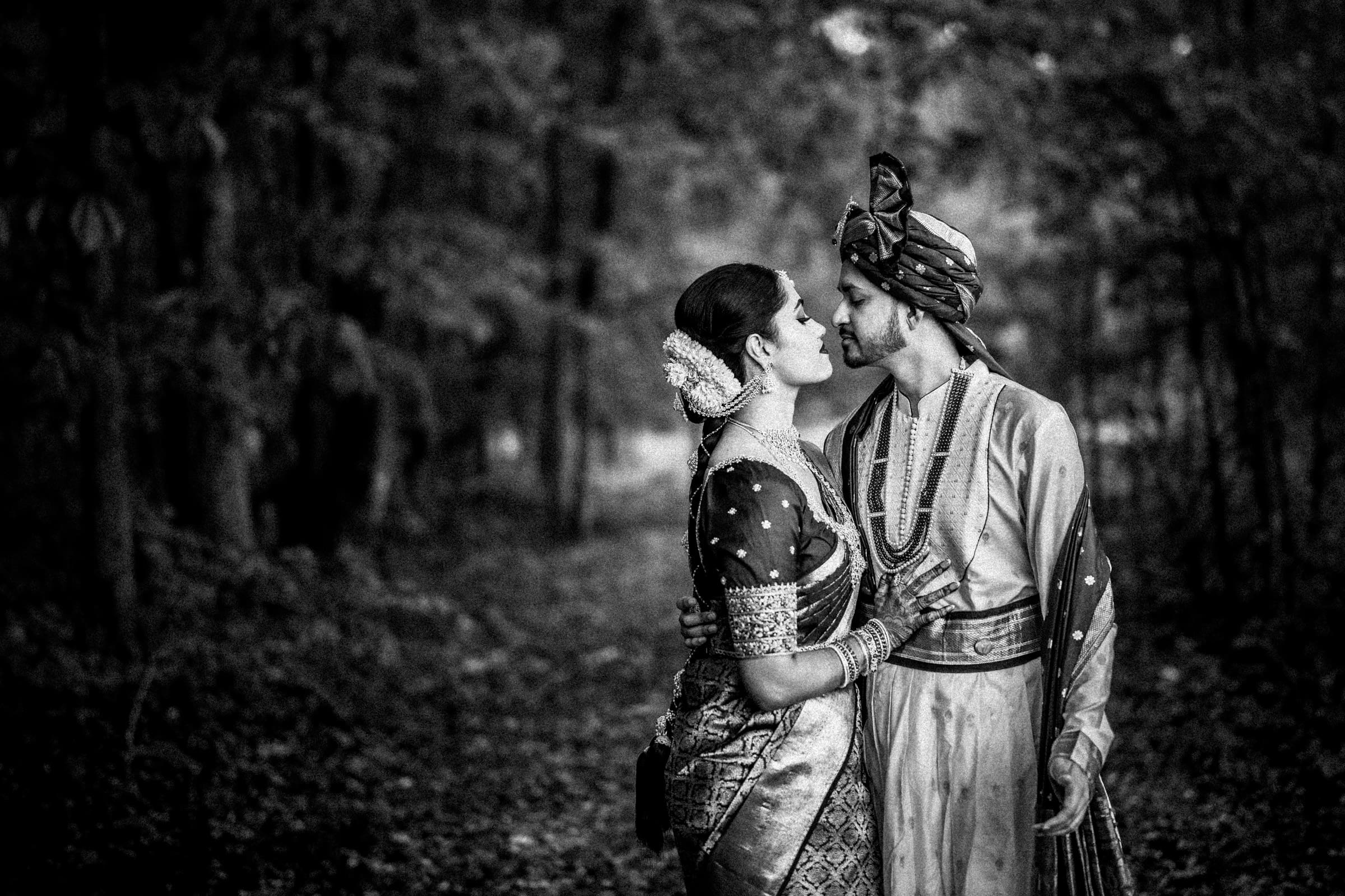 A couple dressed in traditional Indian attire stand closely together in a forest, gazing lovingly into each other’s eyes. The photo is black and white, with blurred trees in the background.