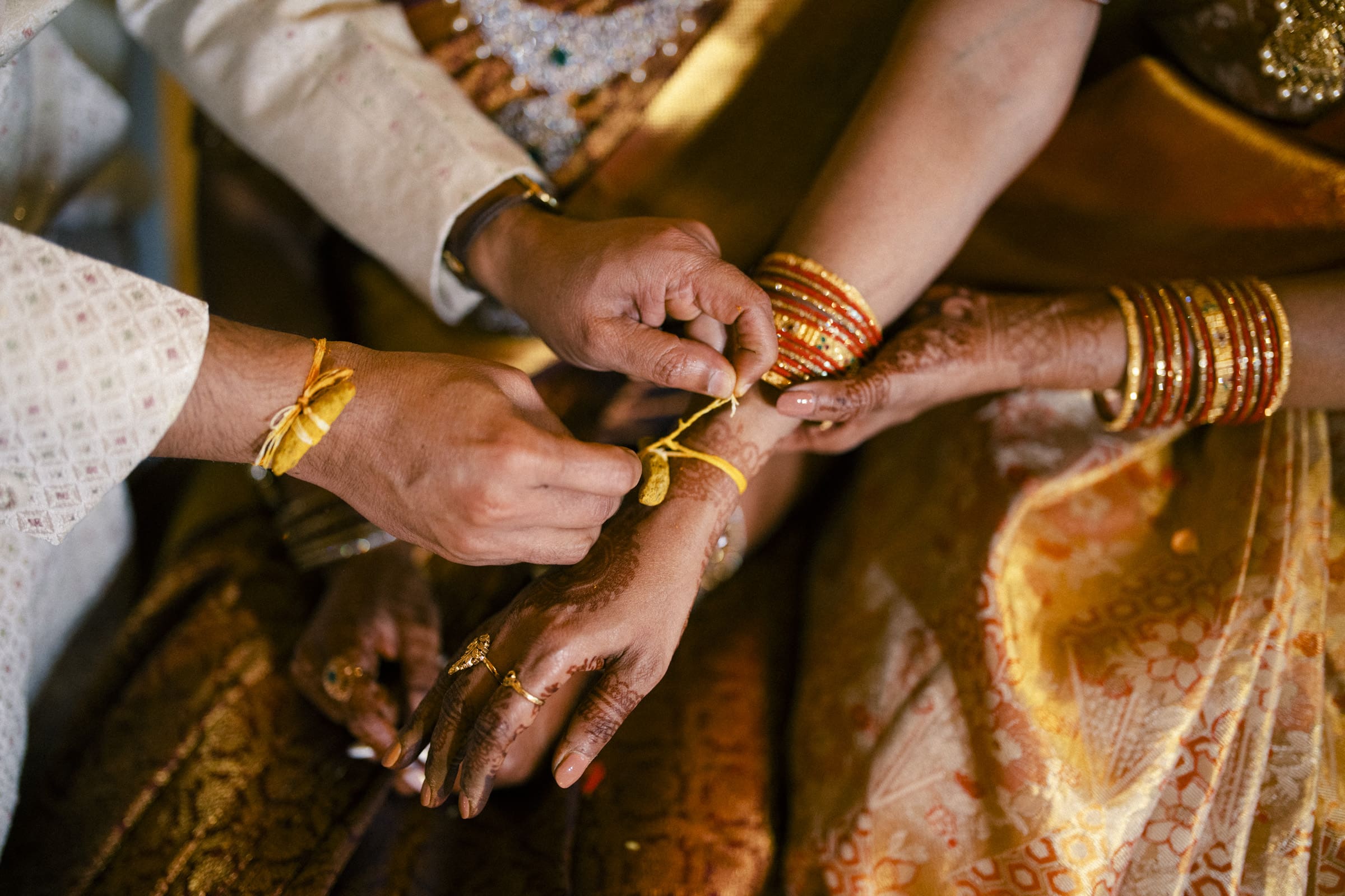 Close-up of hands tying a yellow sacred thread around a womans wrist during a traditional Indian ceremony. She wears gold bangles and a gold saree, with henna designs on her hands.