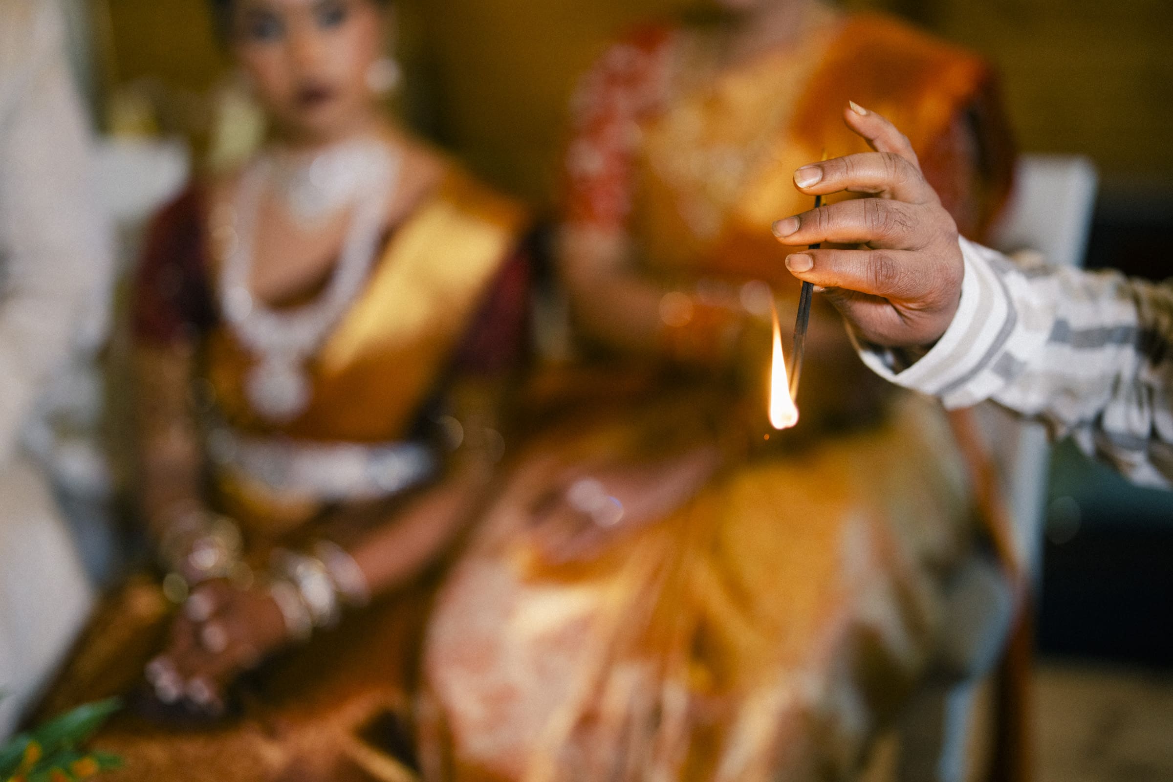 A close-up of a hand holding a small flame, with two women in traditional Indian attire and jewelry blurred in the background, suggesting a ceremonial or ritual setting.