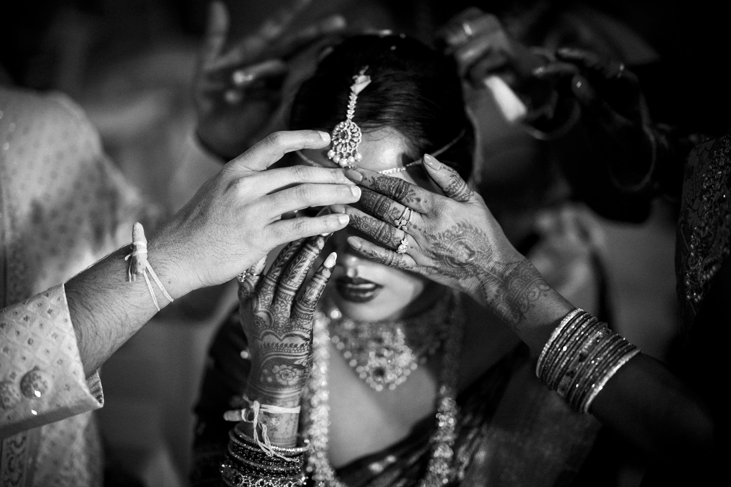 A black-and-white photo of a bride with intricate henna on her hands and ornate jewelry, as several hands gently cover her eyes during a wedding ceremony.