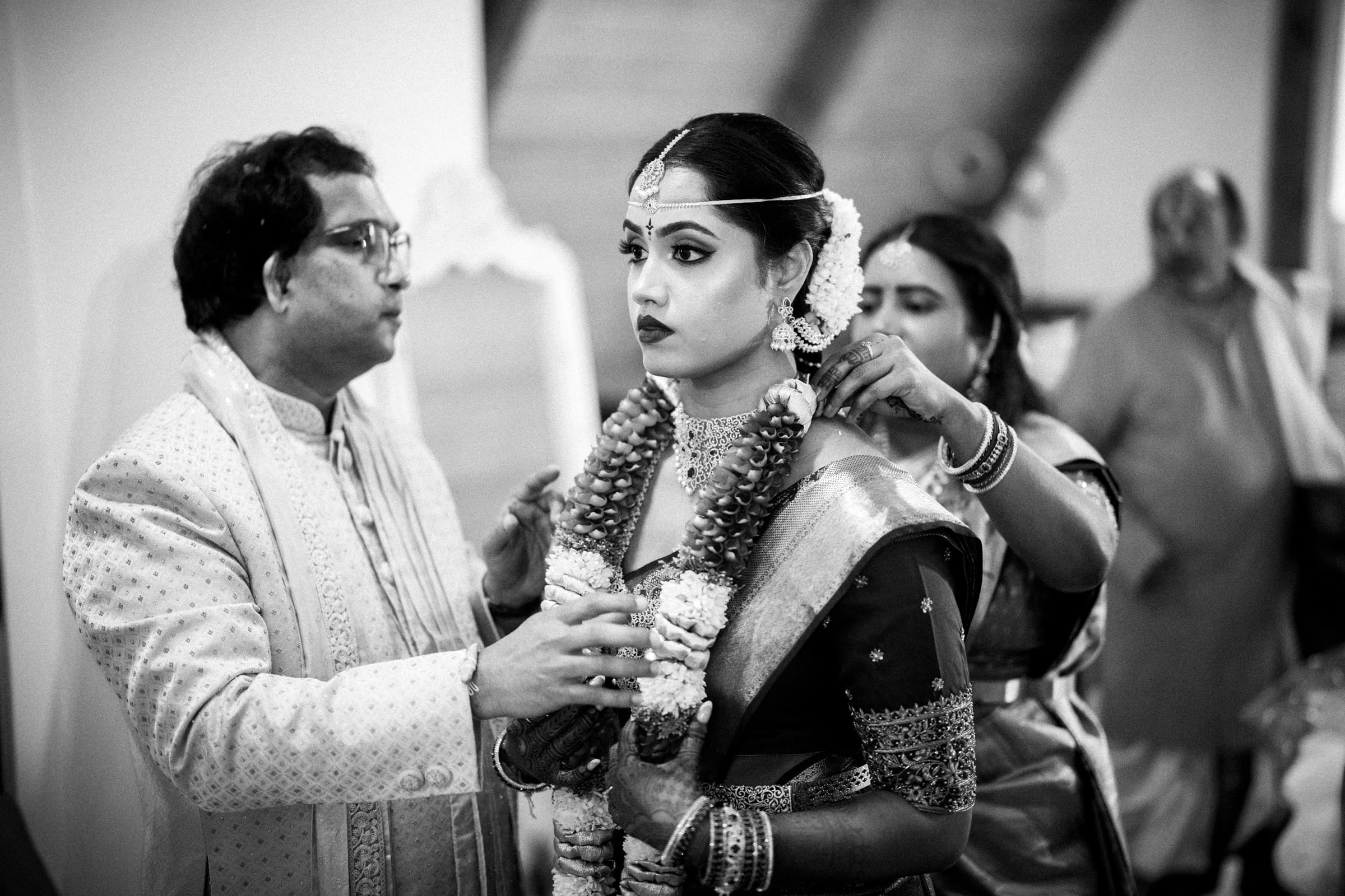 A bride in traditional Indian attire and jewelry stands as a man and woman help place floral garlands around her neck during a wedding ceremony. The scene is captured in black and white.