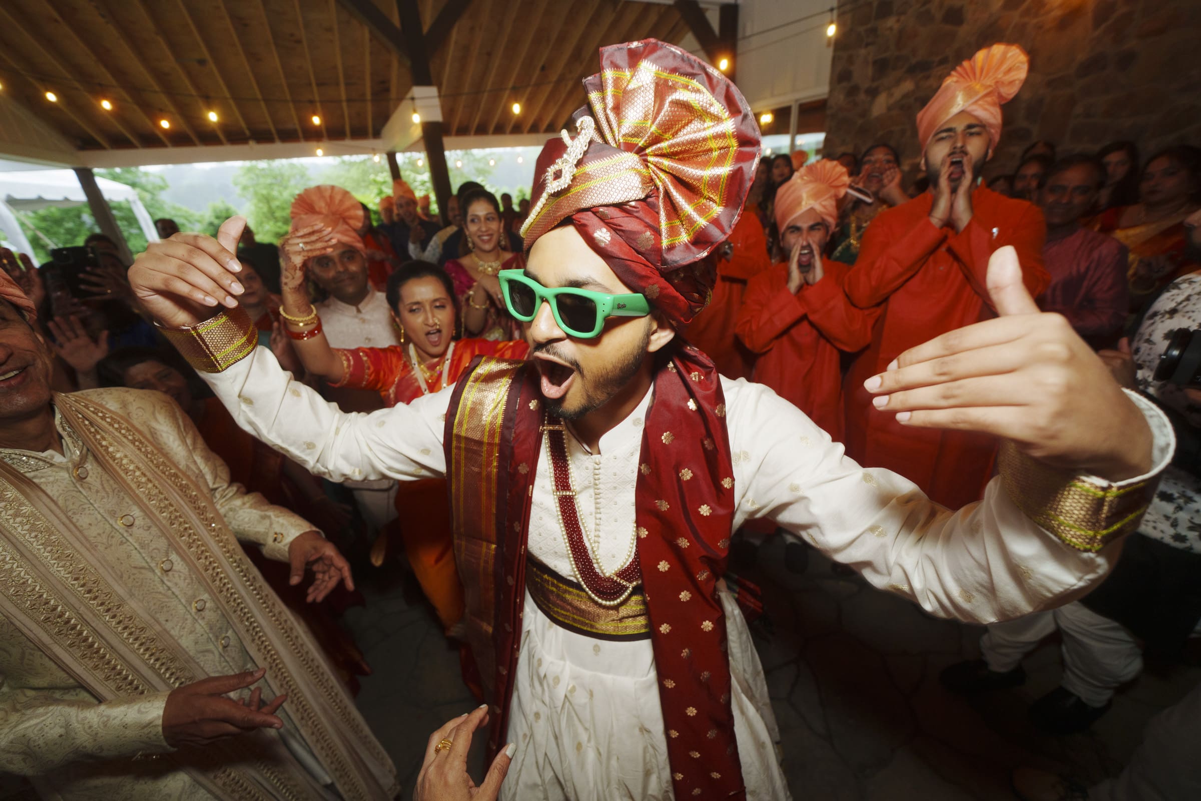A groom wearing a red turban and sunglasses dances joyfully through an energetic crowd during his Baraat at The Pavilion at Carriage Farm.