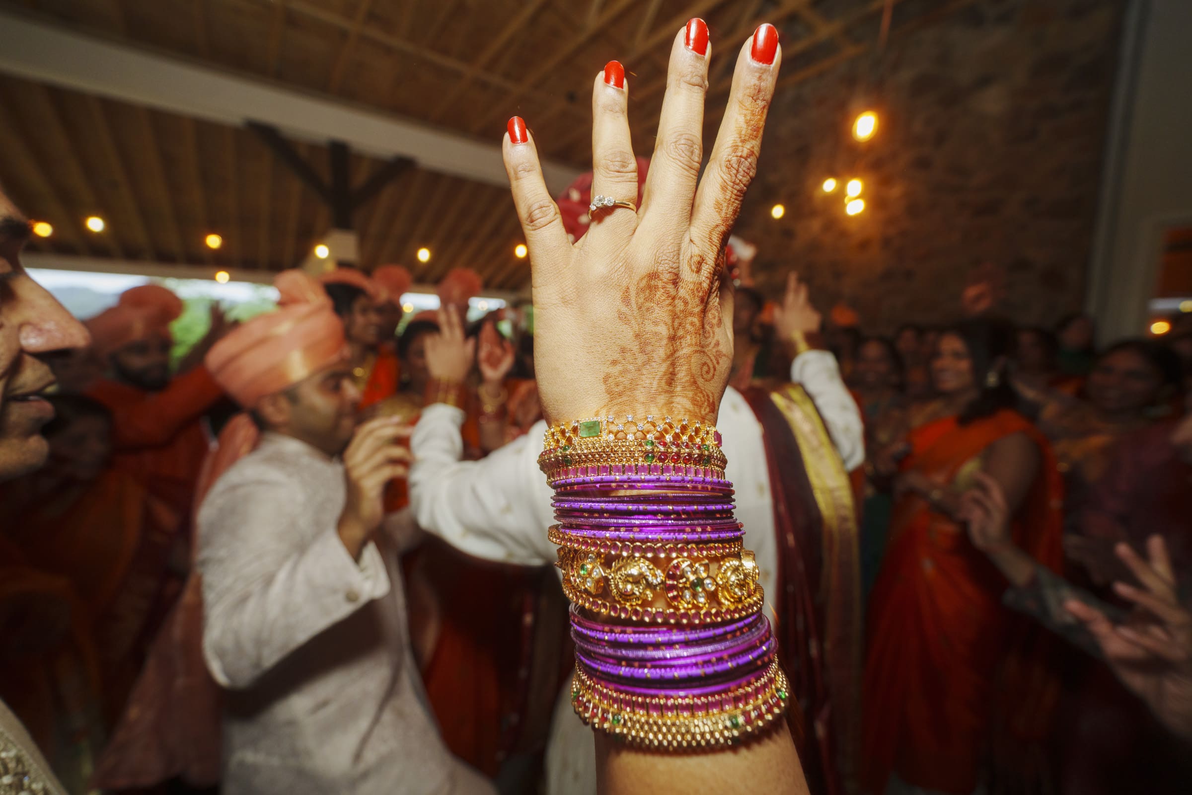 A close-up of a woman's raised hand featuring detailed mehndi designs and vibrant purple bangles, with wedding guests dancing in the background at Carriage Farm.