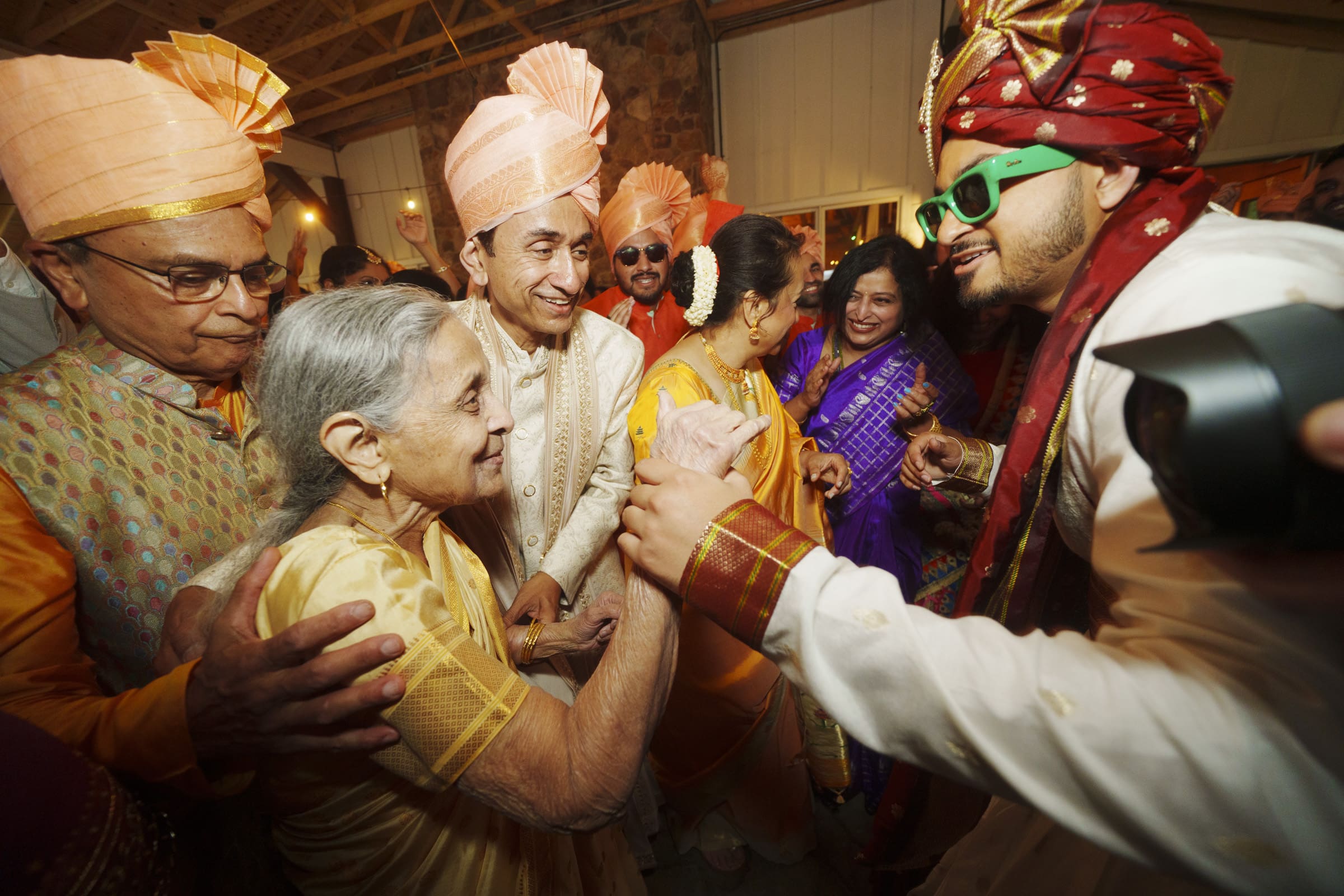 A joyful group at an Indian wedding, with men in traditional attire and turbans, an elderly woman in yellow greeting a groom in a red turban and sunglasses, surrounded by smiling guests in colorful clothing.