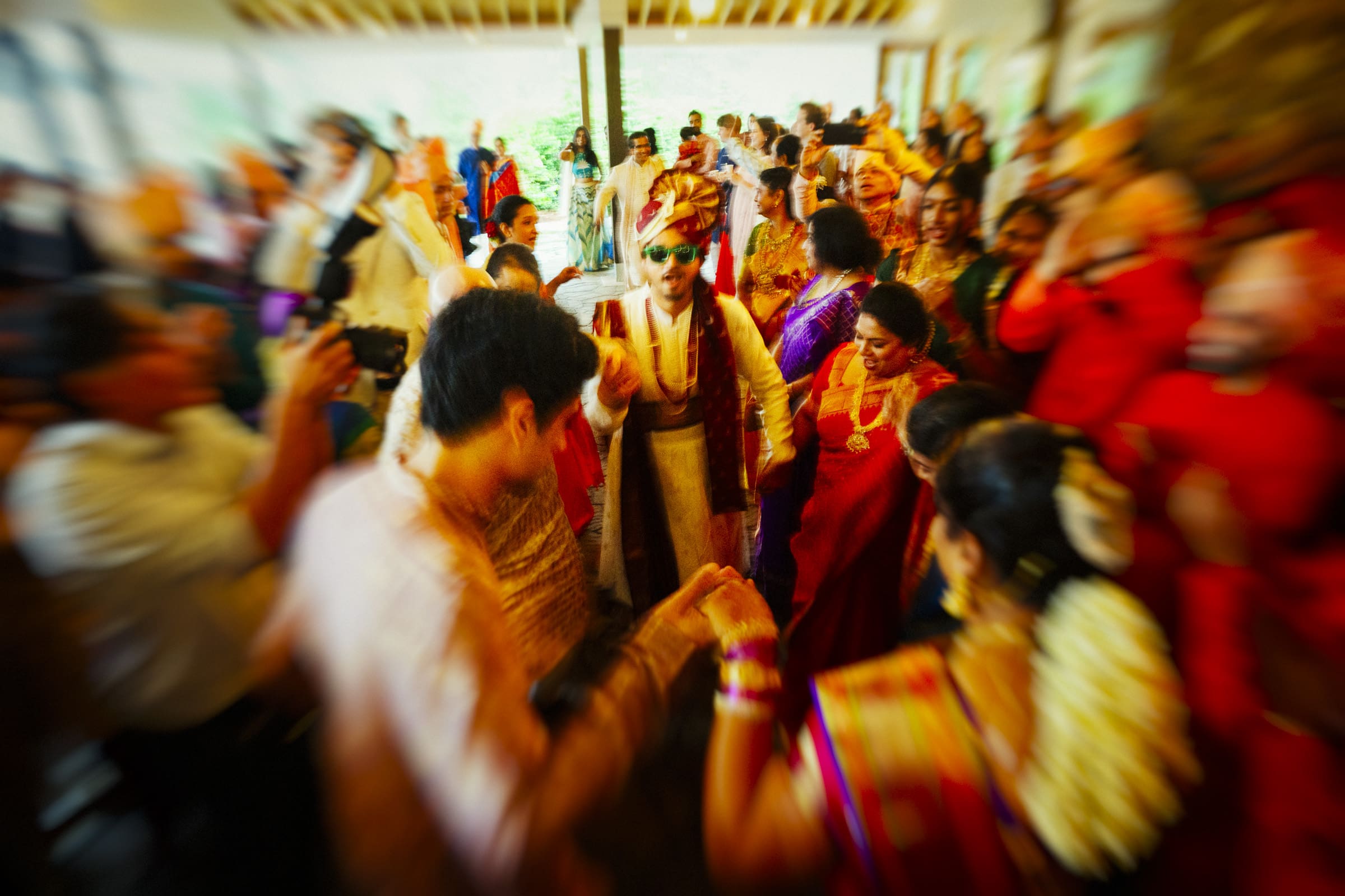 A groom in traditional Indian attire and sunglasses dances joyfully during his Baraat celebration at The Pavilion at Carriage Farm.