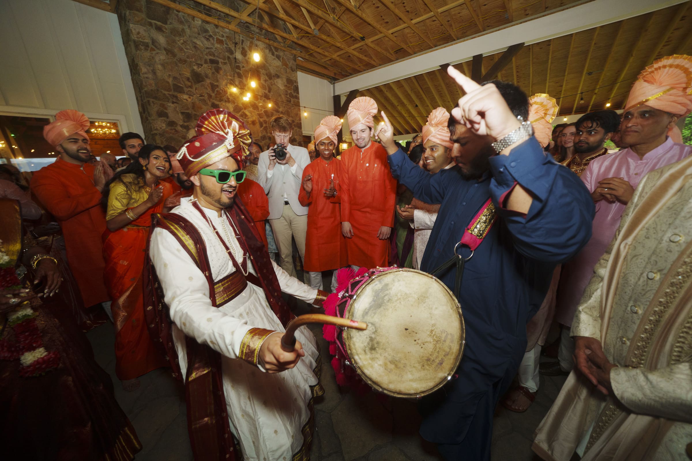 A lively group of wedding guests dance energetically indoors while the groom plays the dhol drum at a Raleigh Indian wedding celebration.