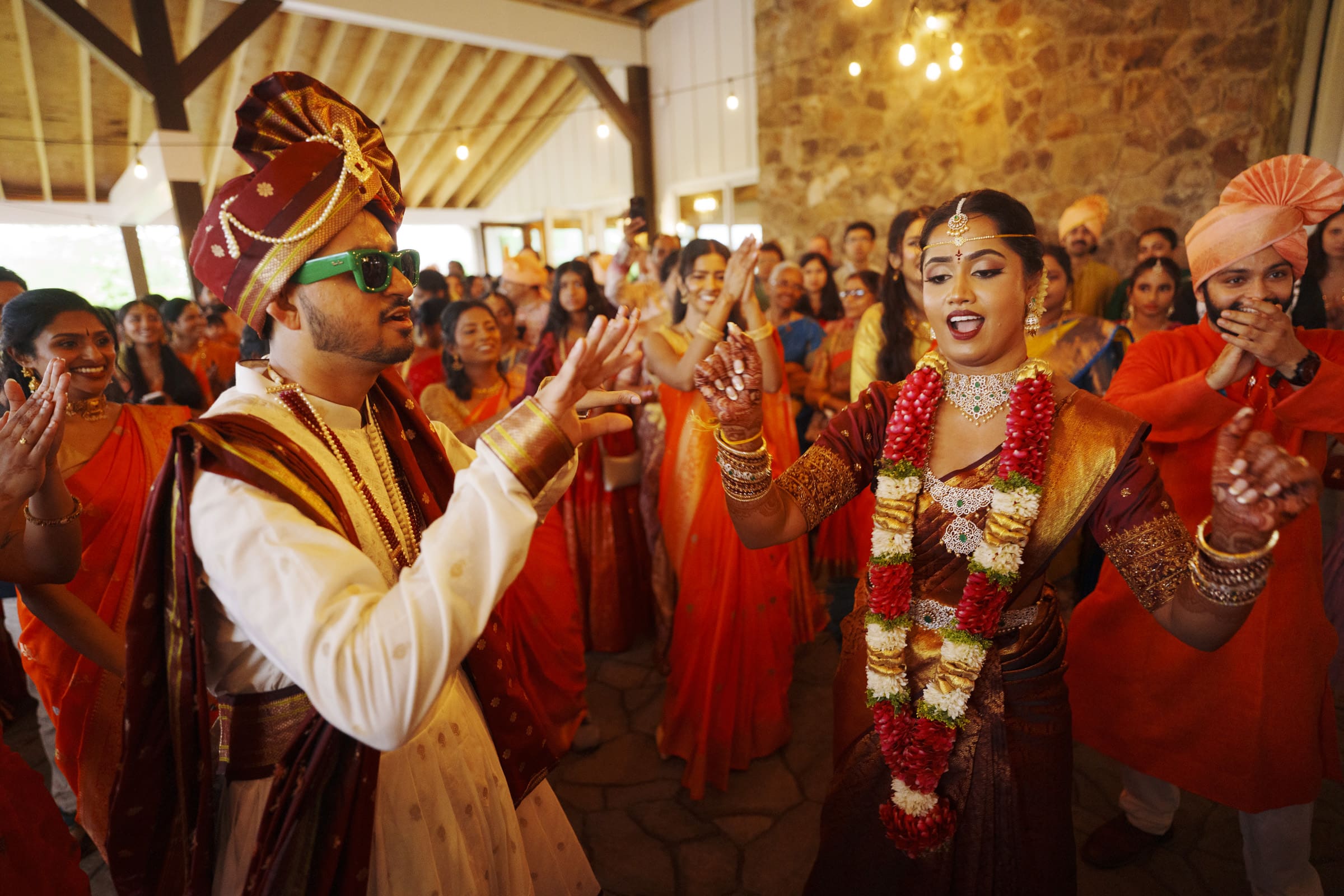 A bride and groom joyfully dance together surrounded by a cheering crowd of family and friends during a lively outdoor Baraat at The Pavilion at Carriage Farm.