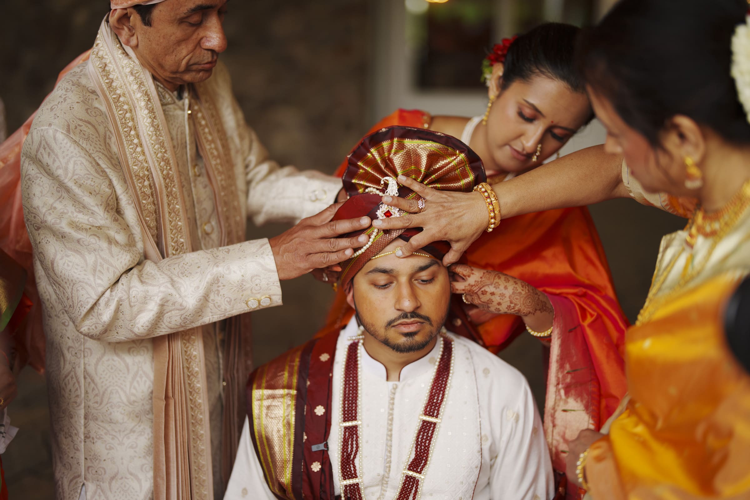 Family members carefully adjust a groom's ornate red turban during a traditional ceremonial ritual at The Pavilion at Carriage Farm.