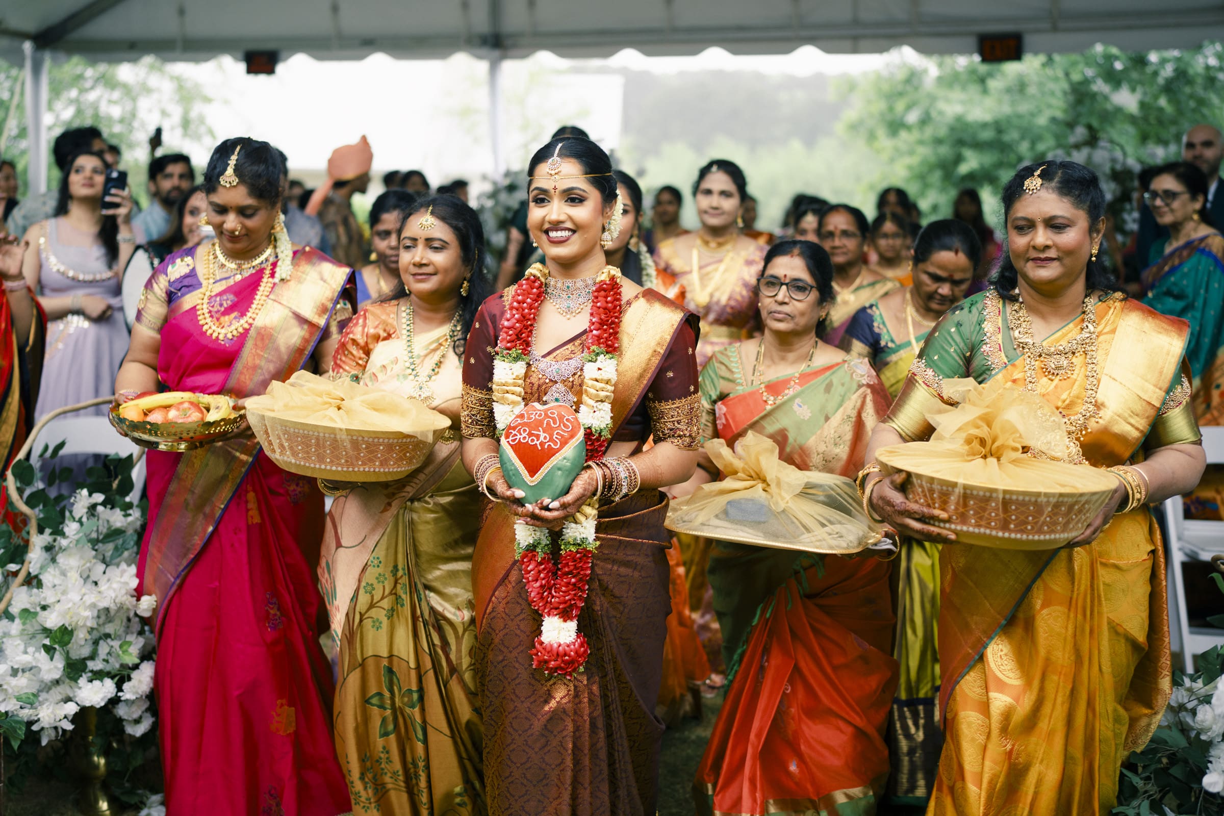 A radiant Indian bride carries a ceremonial coconut as she walks down the aisle during her wedding ceremony entrance at The Pavilion at Carriage Farm.