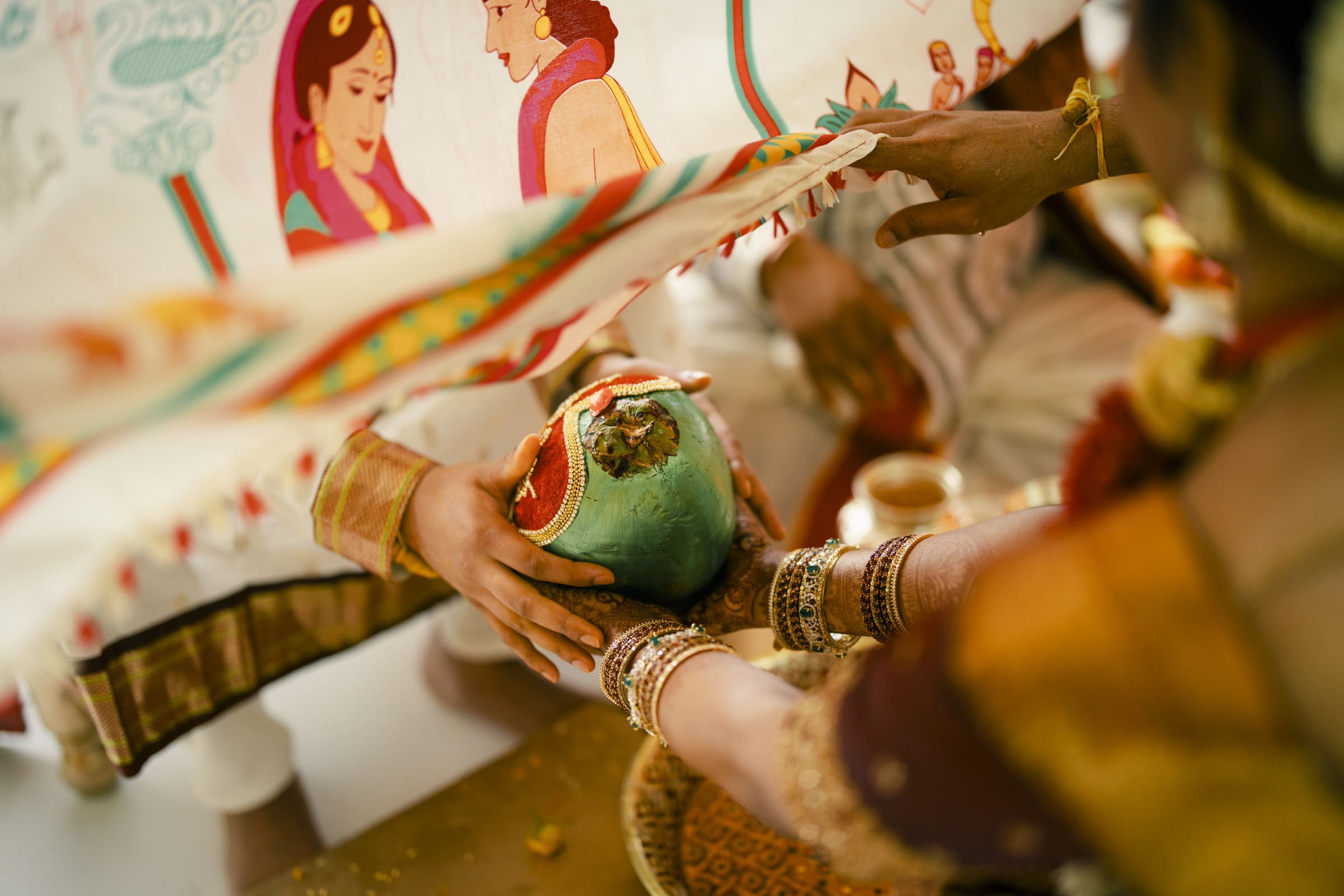 Two people in traditional Indian attire hold a decorated coconut together during a wedding ritual, with a colorful cloth featuring illustrated faces hanging above their hands.