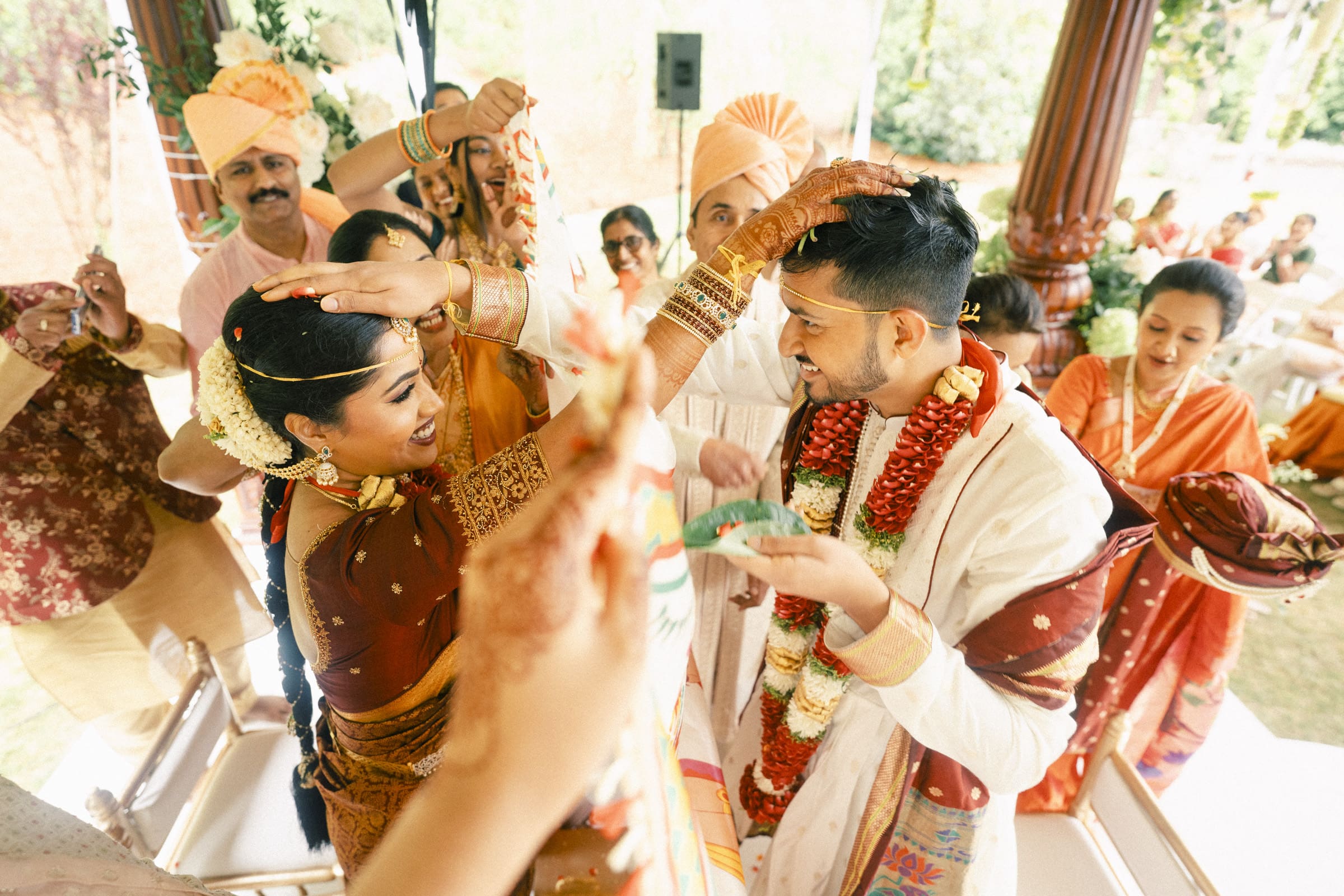 A bride and groom in traditional Indian attire playfully touch each other’s heads during a wedding ceremony, surrounded by joyful family and friends dressed in colorful outfits.