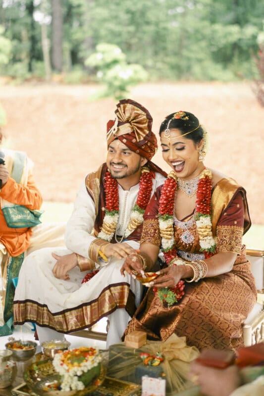 A smiling couple in traditional Indian wedding attire sits together, adorned with flower garlands and jewelry, participating in a wedding ceremony with decorative items in front of them.