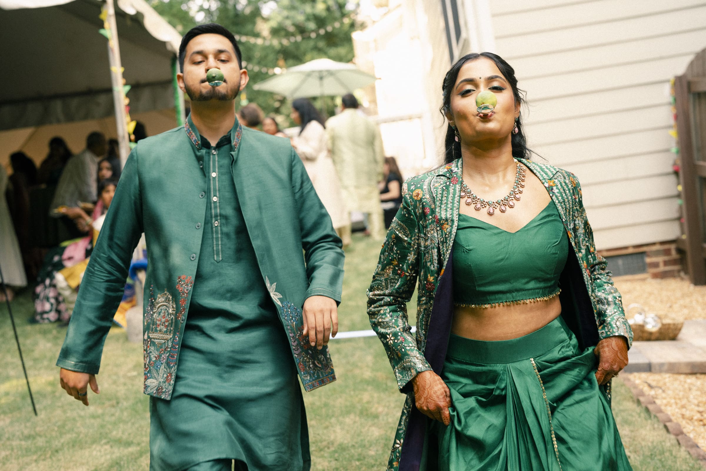 Two people wearing matching green traditional outfits walk outdoors with limes held in their mouths, participating in a fun activity at a festive event. Other guests and decorations are visible in the background.