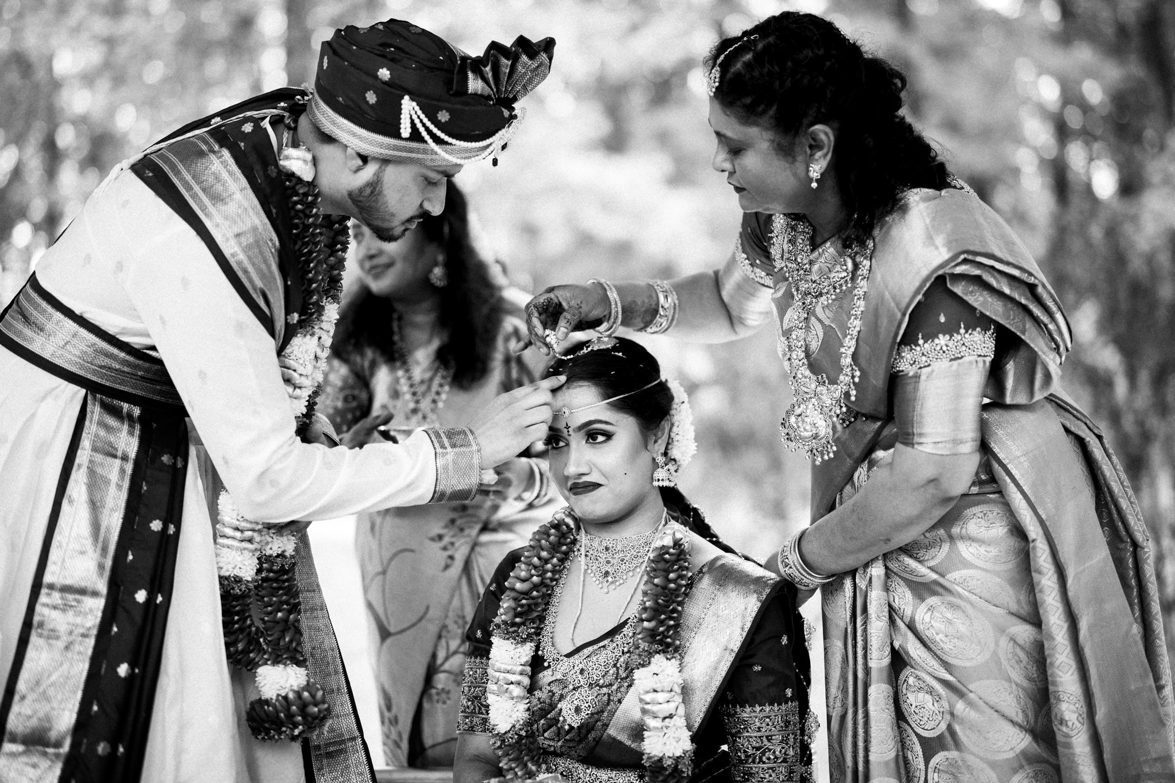 A bride sits while a groom and another woman, both in traditional attire, perform a wedding ritual by placing something on her head. The scene is outdoors and in black and white.