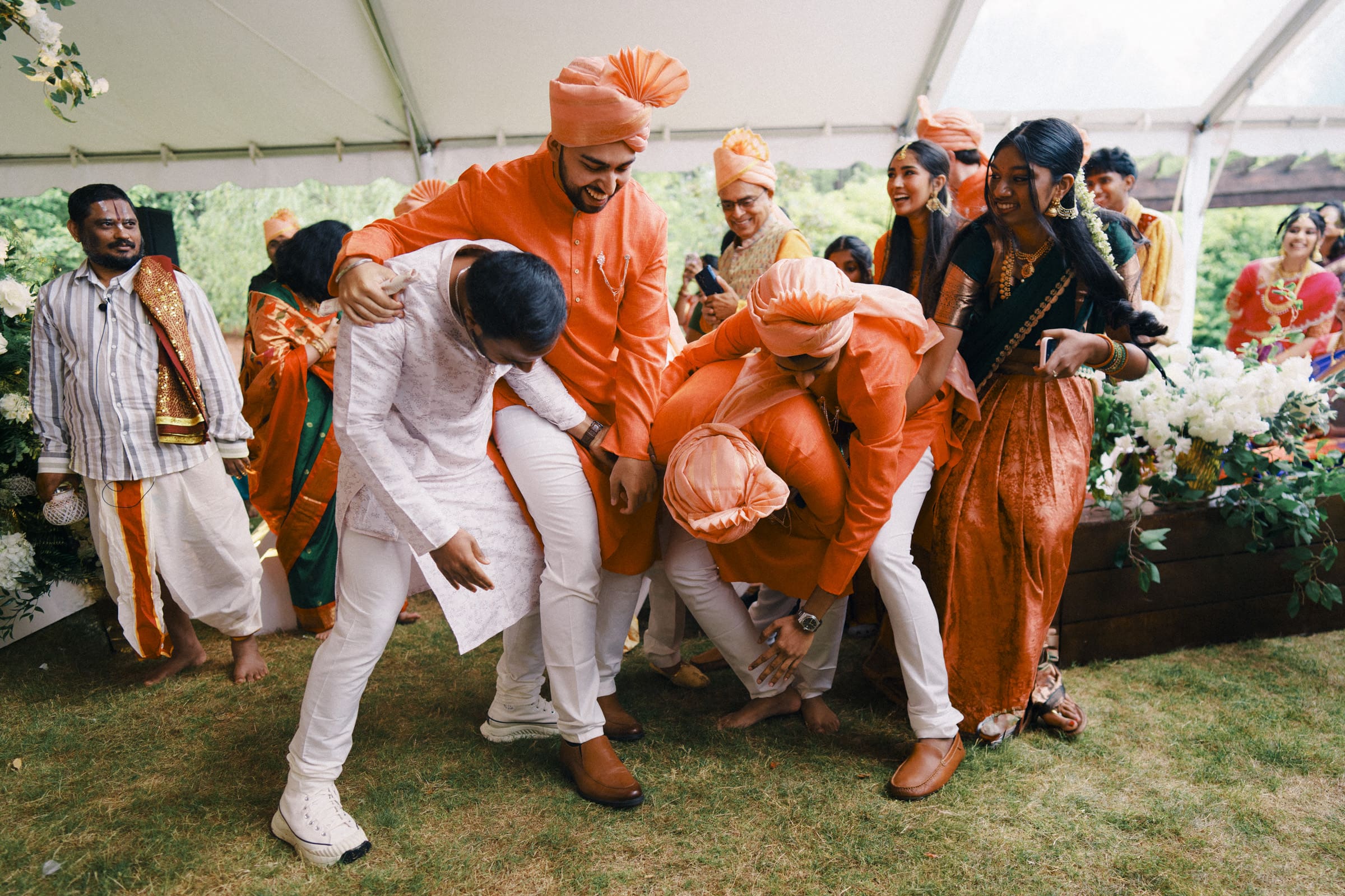 A joyful group of people, mostly dressed in bright orange traditional outfits, laugh and tussle playfully as they look for the groom's shoe at an outdoor celebration under a white tent, surrounded by greenery and flowers at a traditional South Indian wedding ceremony in Raleigh.
