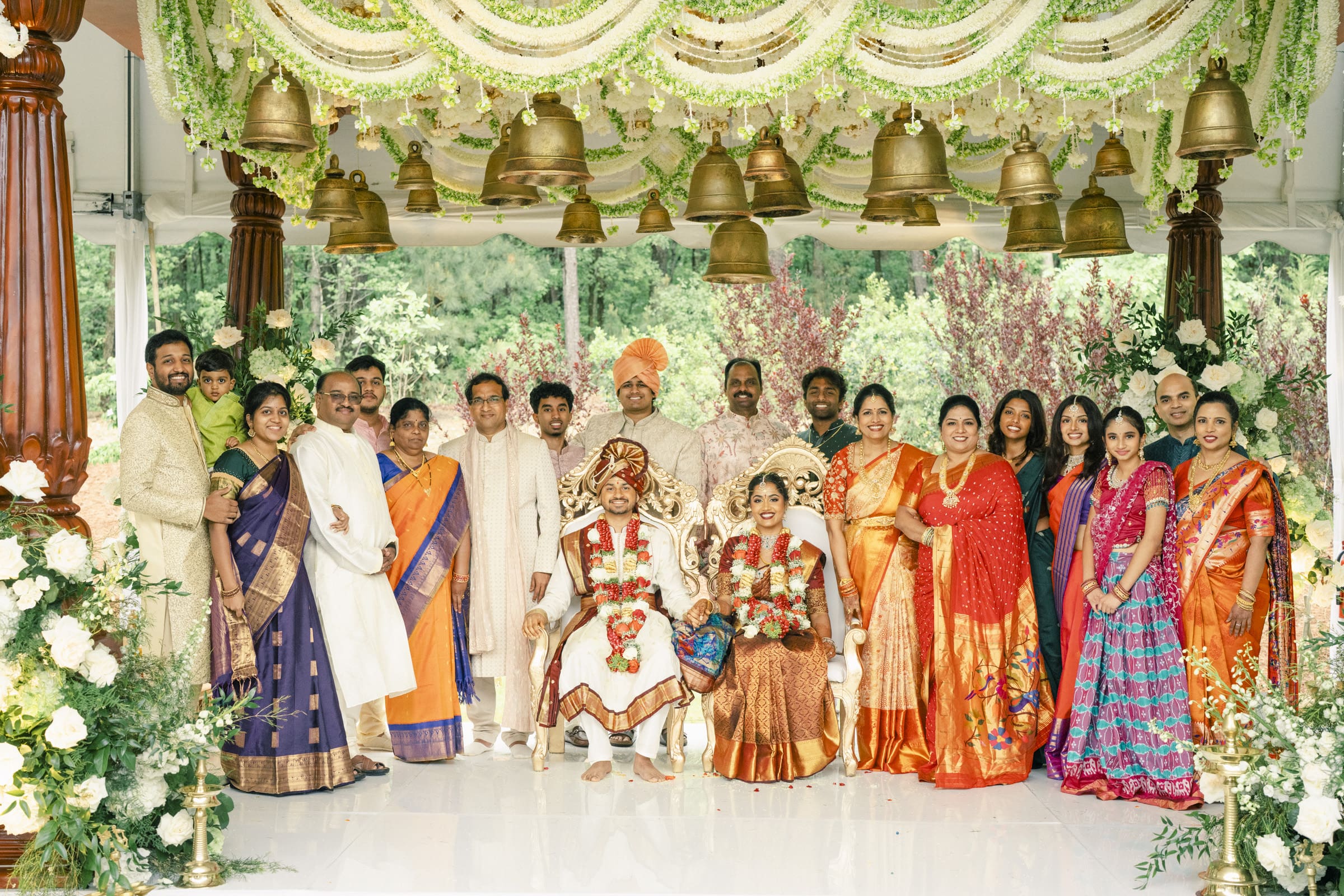 A large group poses at an Indian wedding ceremony, with the bride and groom seated at the center in traditional attire, surrounded by family and friends in colorful outfits, under a decorated canopy with bells and greenery.