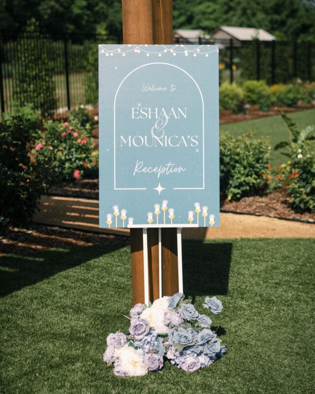 A blue welcome sign reading “Eshaan & Mounica’s Reception” is displayed outdoors on a wooden stand, surrounded by decorative white and lavender flowers and lush garden greenery in the background.