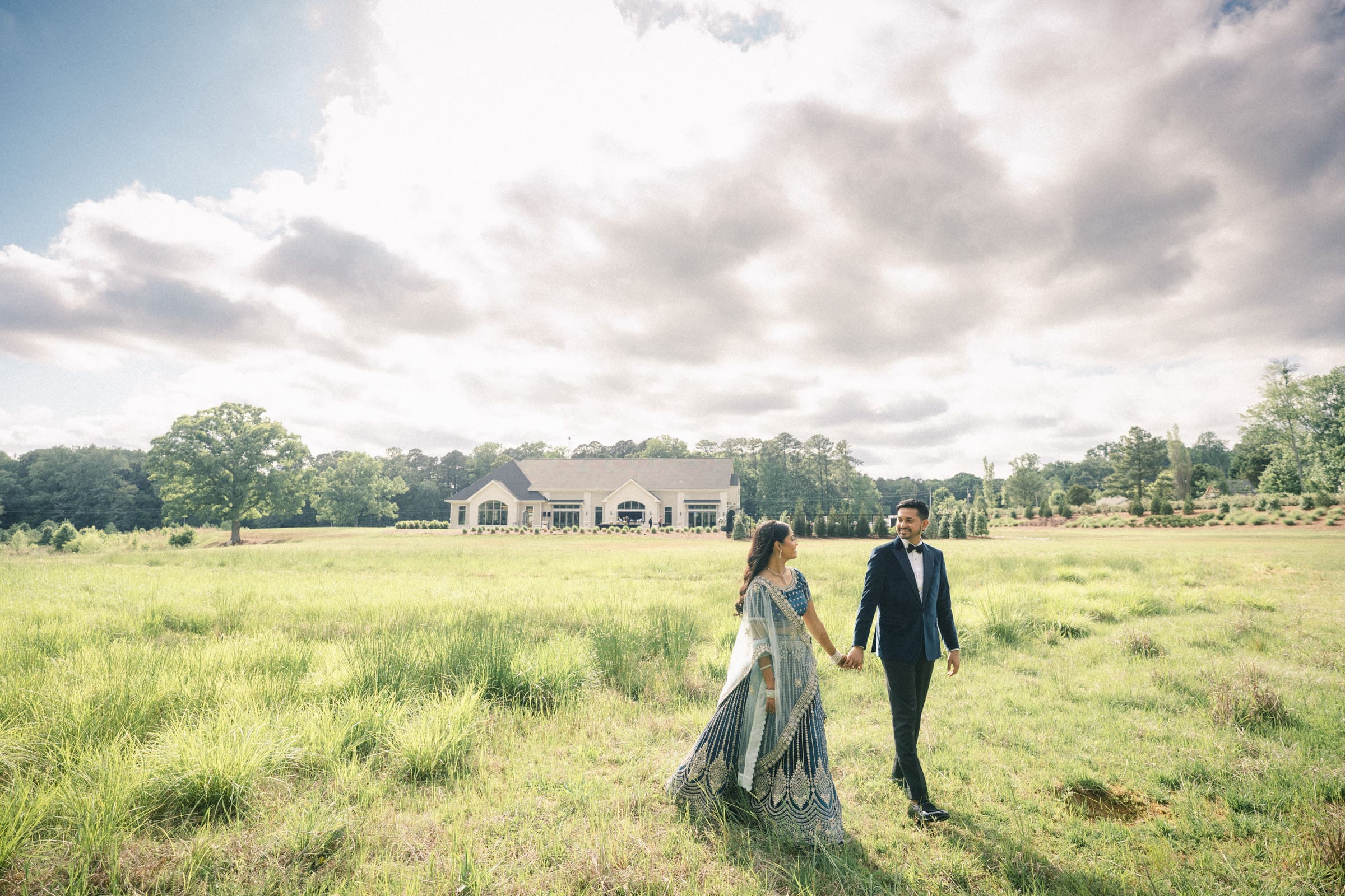 A bride and groom walk hand-in-hand across a sunlit grassy field with the white historic estate house in the background at Donovan Manor.